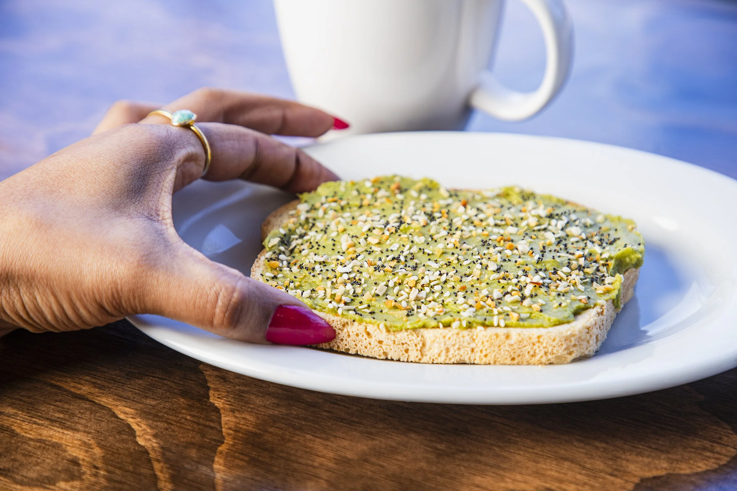 A person with red nail polish and a gold ring on their finger is holding a white plate with a slice of bread spread with green avocado and topped with mixed seeds and herbs. There is a white mug in the background on a wooden table.