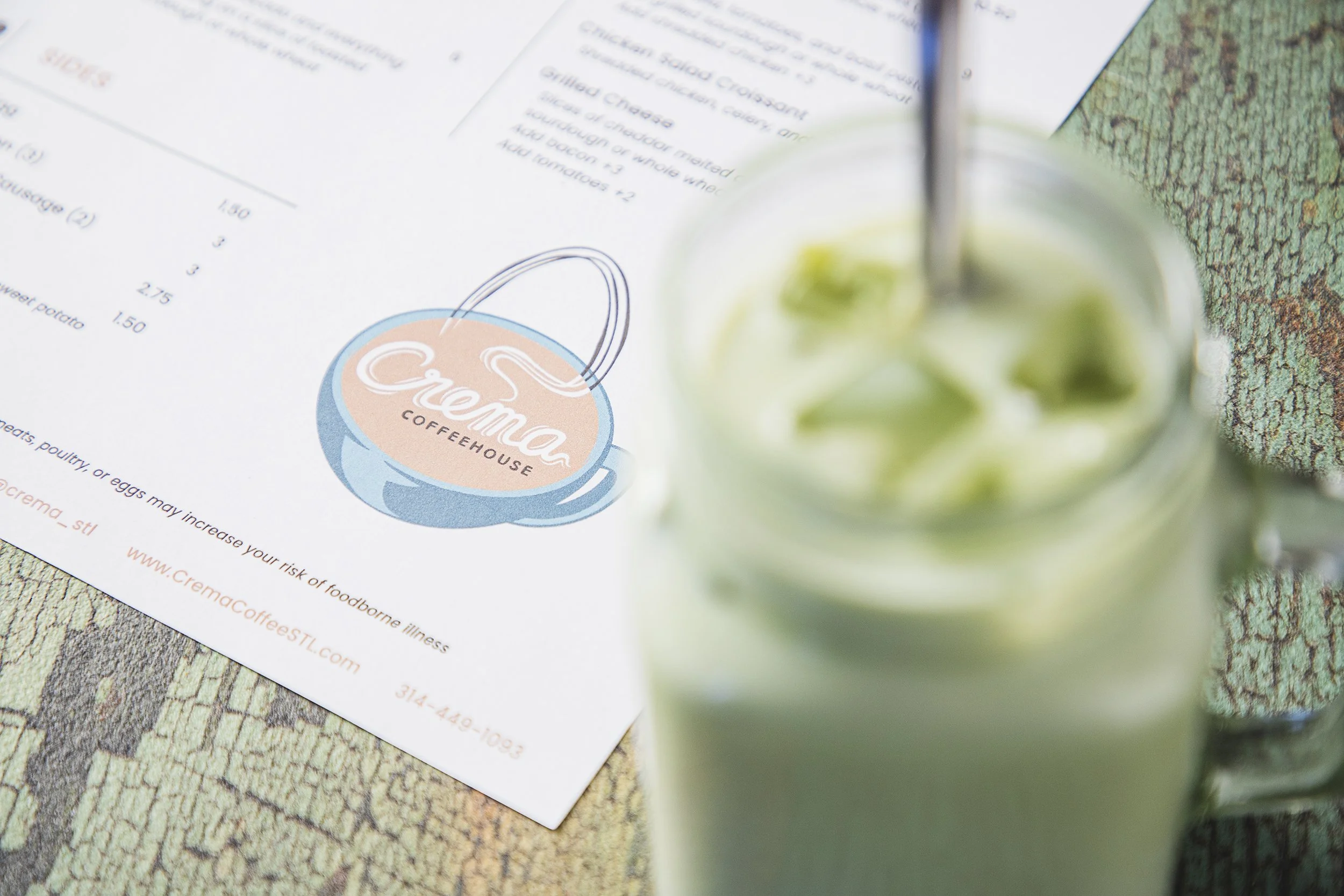 A menu from Crema Coffeehouse is on a textured surface, partially blurred, with a mason jar of green beverage and a metal straw in the foreground.