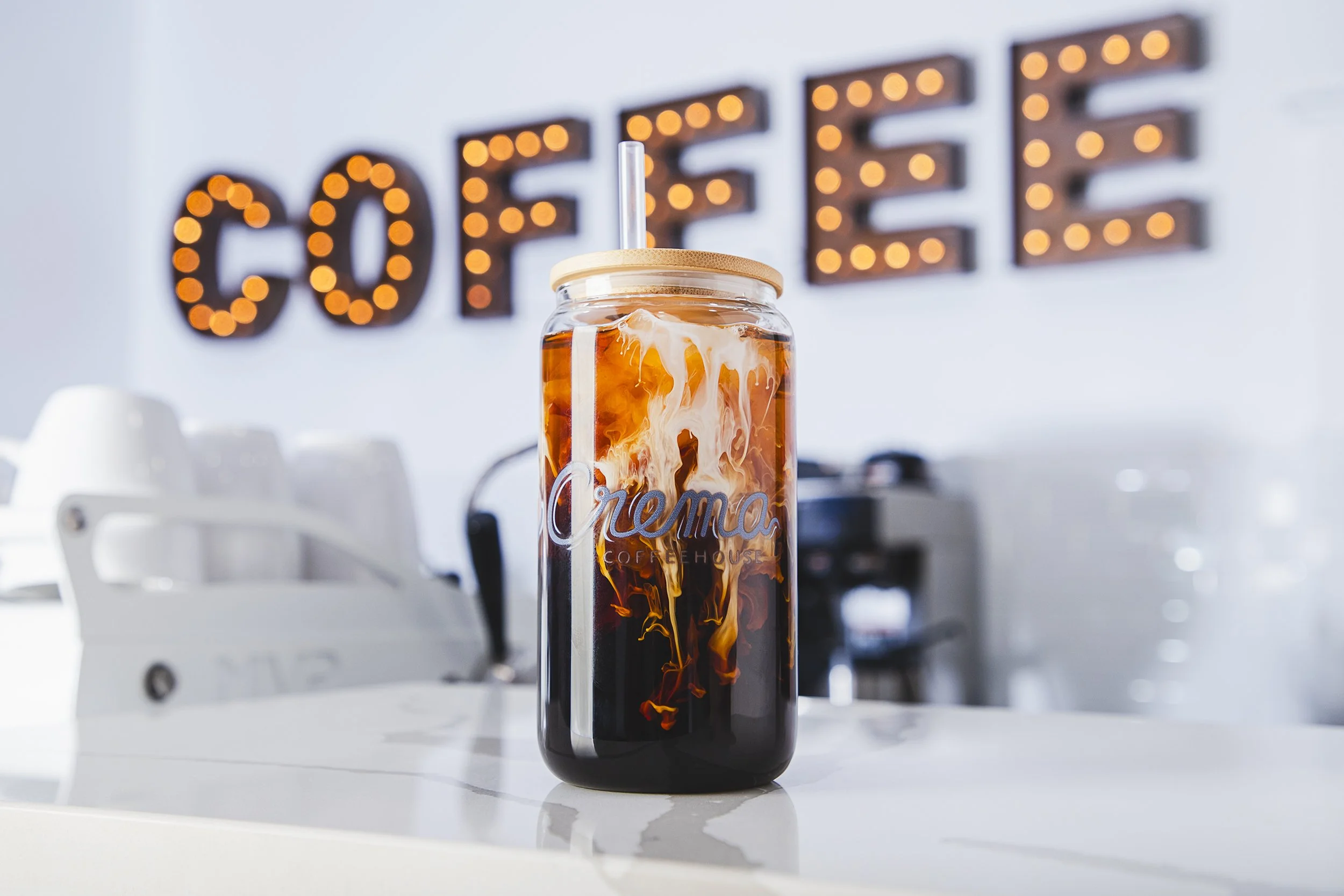 A jar filled with iced coffee and cream with a straw, placed on a white counter in a modern coffee shop with a "COFFEE" sign illuminated in the background.