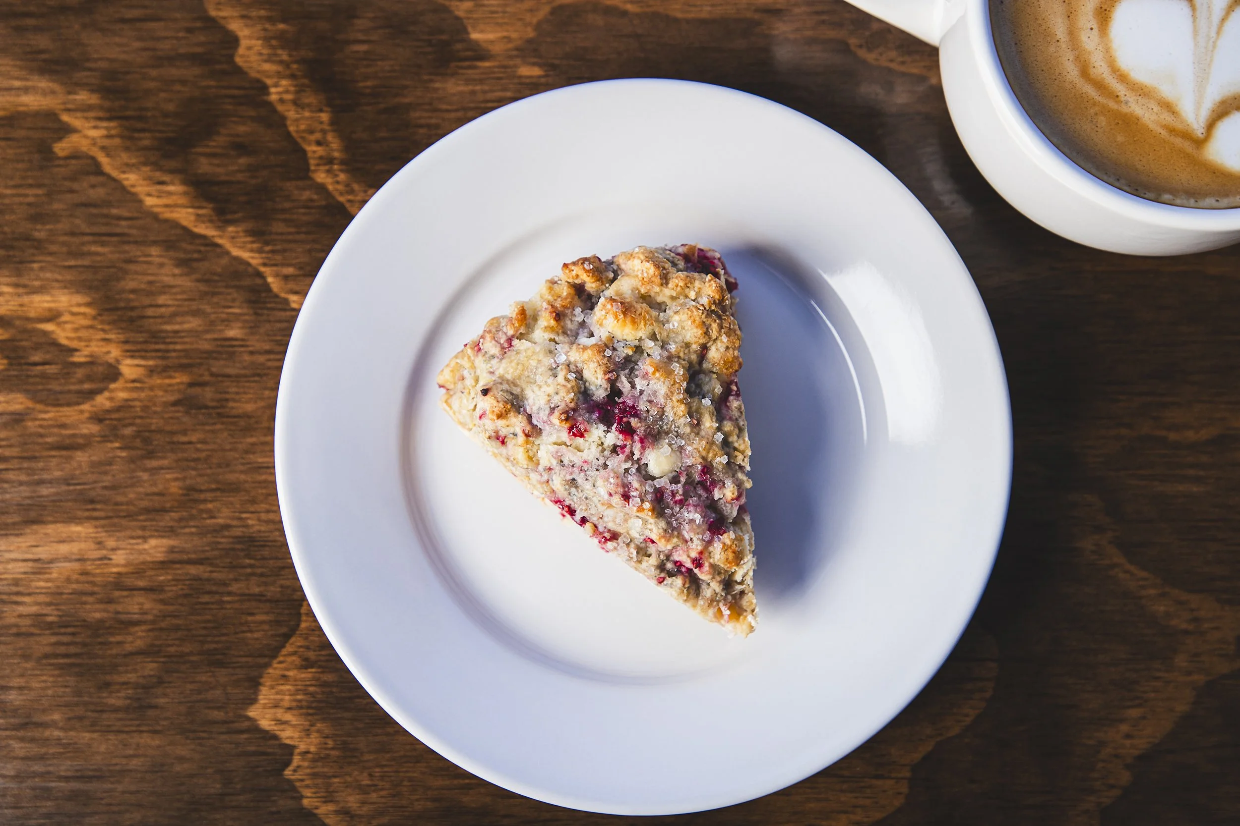 A slice of fruit crumble pie on a white plate, with a cup of coffee with latte art nearby on a wooden table.
