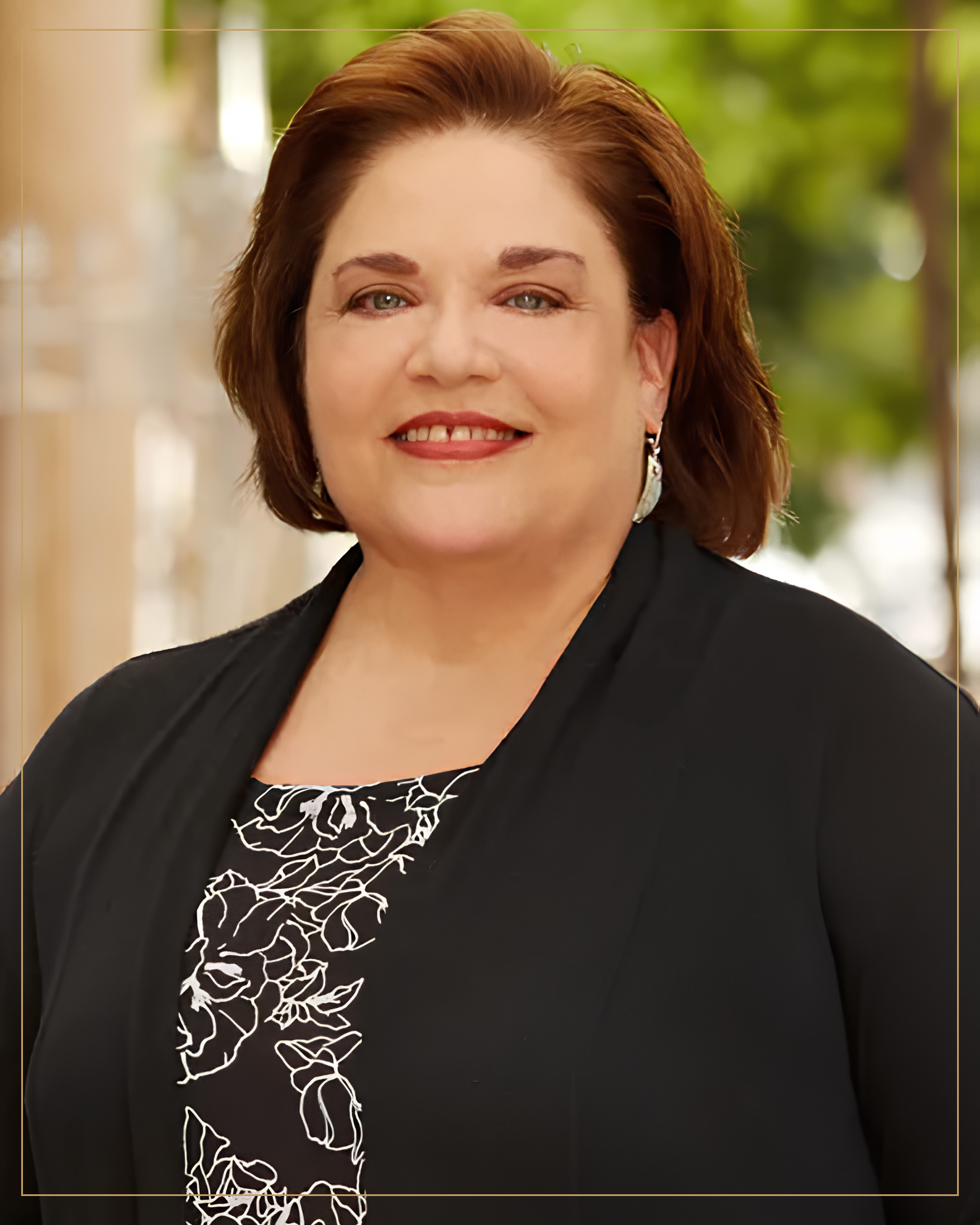 A smiling woman with short, auburn hair, wearing a black blazer and a black and white patterned blouse, standing outdoors with green blurred foliage in the background.