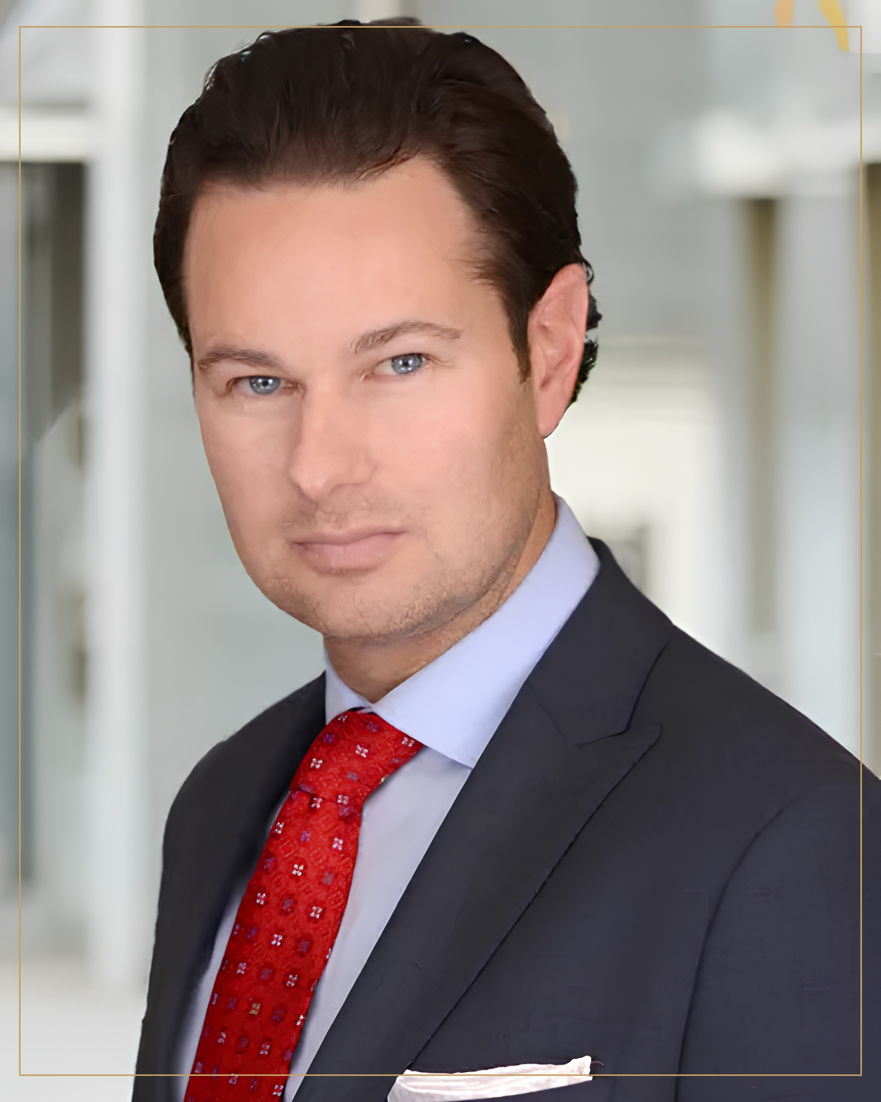 A man in a dark suit, light blue shirt, and red tie, standing in a professional indoor setting.