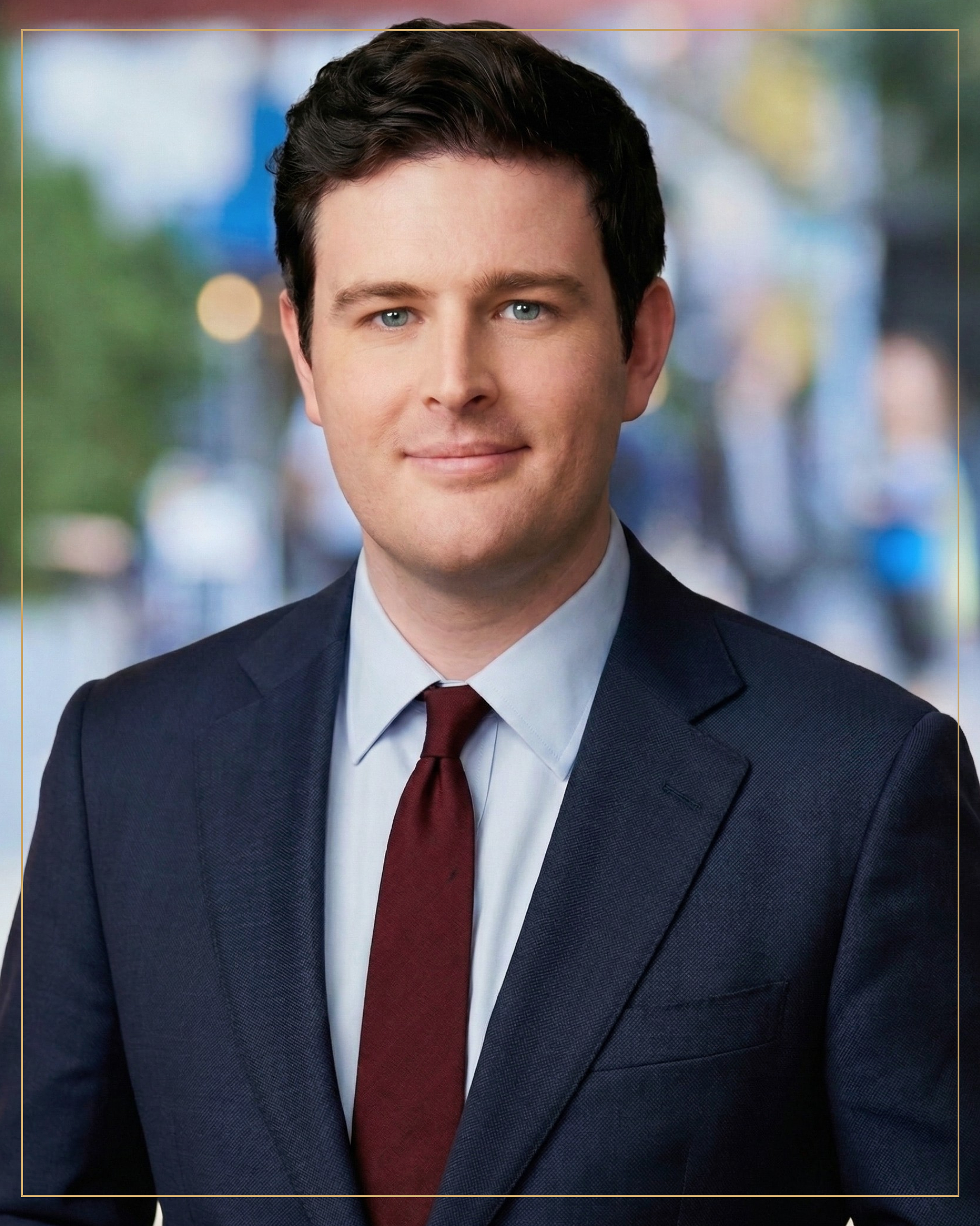 A professional headshot of a young man with dark hair, blue eyes, wearing a navy suit, white shirt, and maroon tie, against a blurred cityscape background.