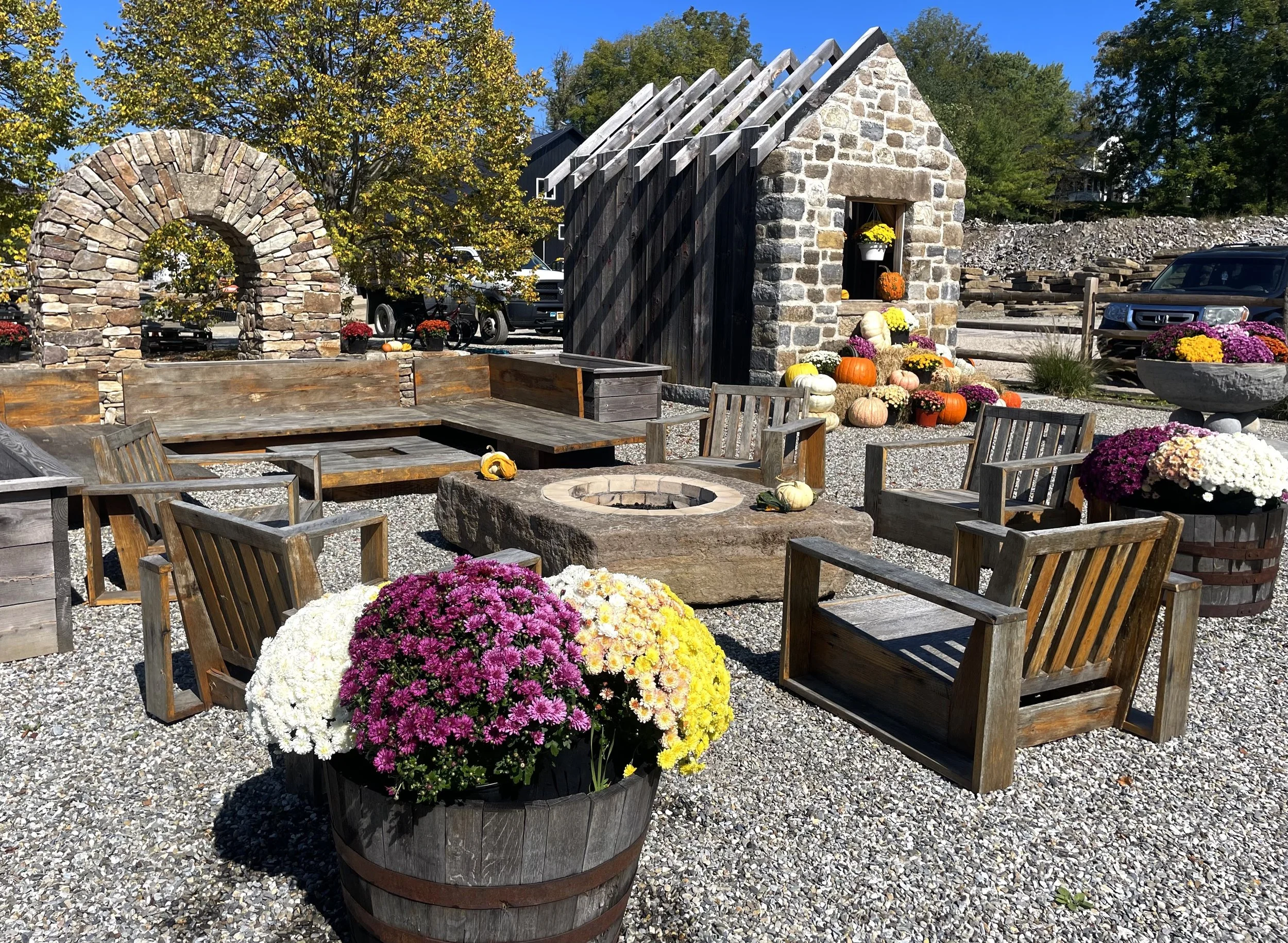 An outdoor seating area with wooden chairs, a stone fire pit, themed fall decorations including pumpkins and chrysanthemums, and a small stone shed, all set on gravel under a clear blue sky.