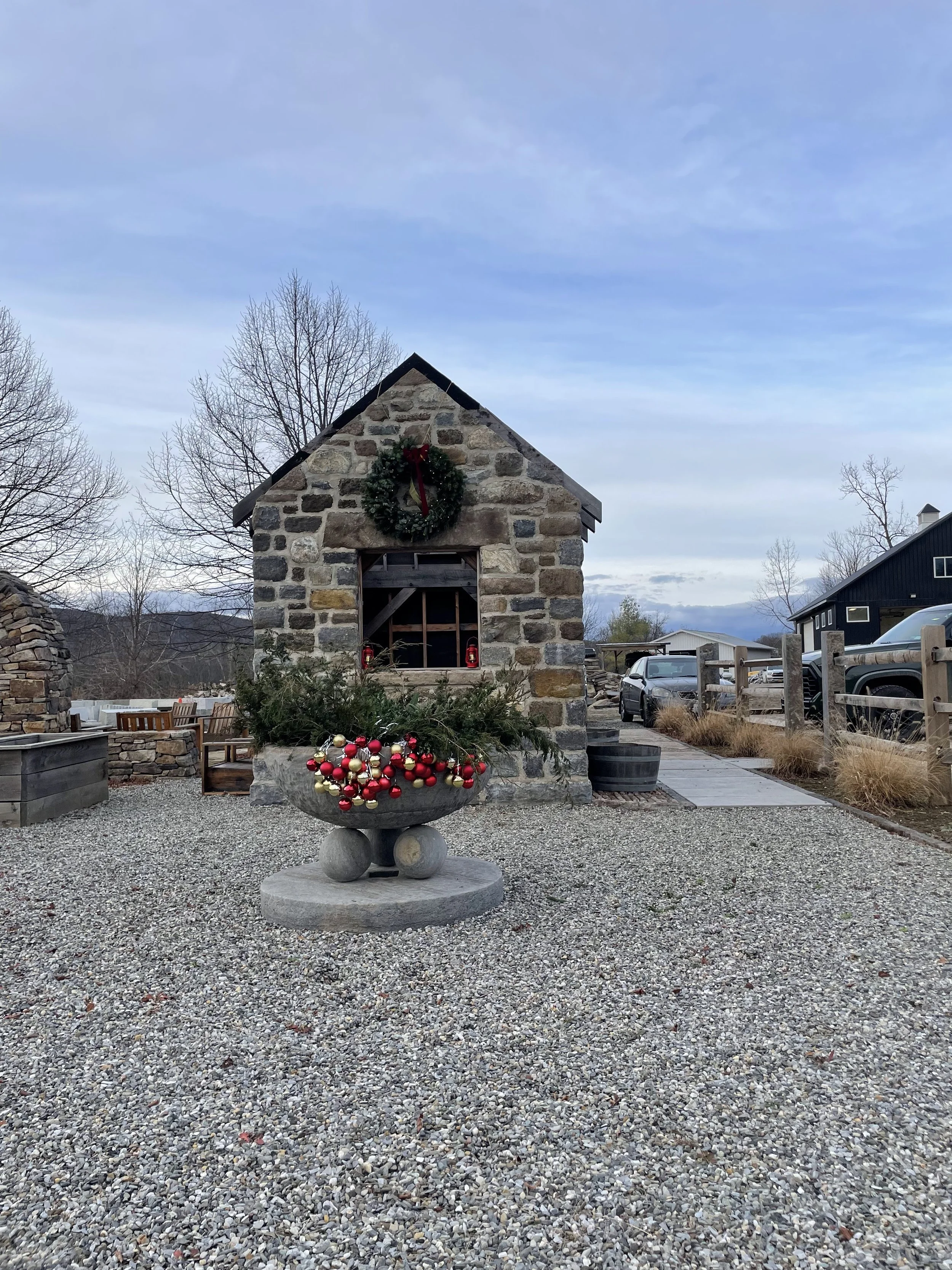 Outdoor scene featuring a small stone building with a Christmas wreath on the roof and a decorative stone basin filled with Christmas ornaments in red, gold, and white, surrounded by a gravel area and wooden benches.