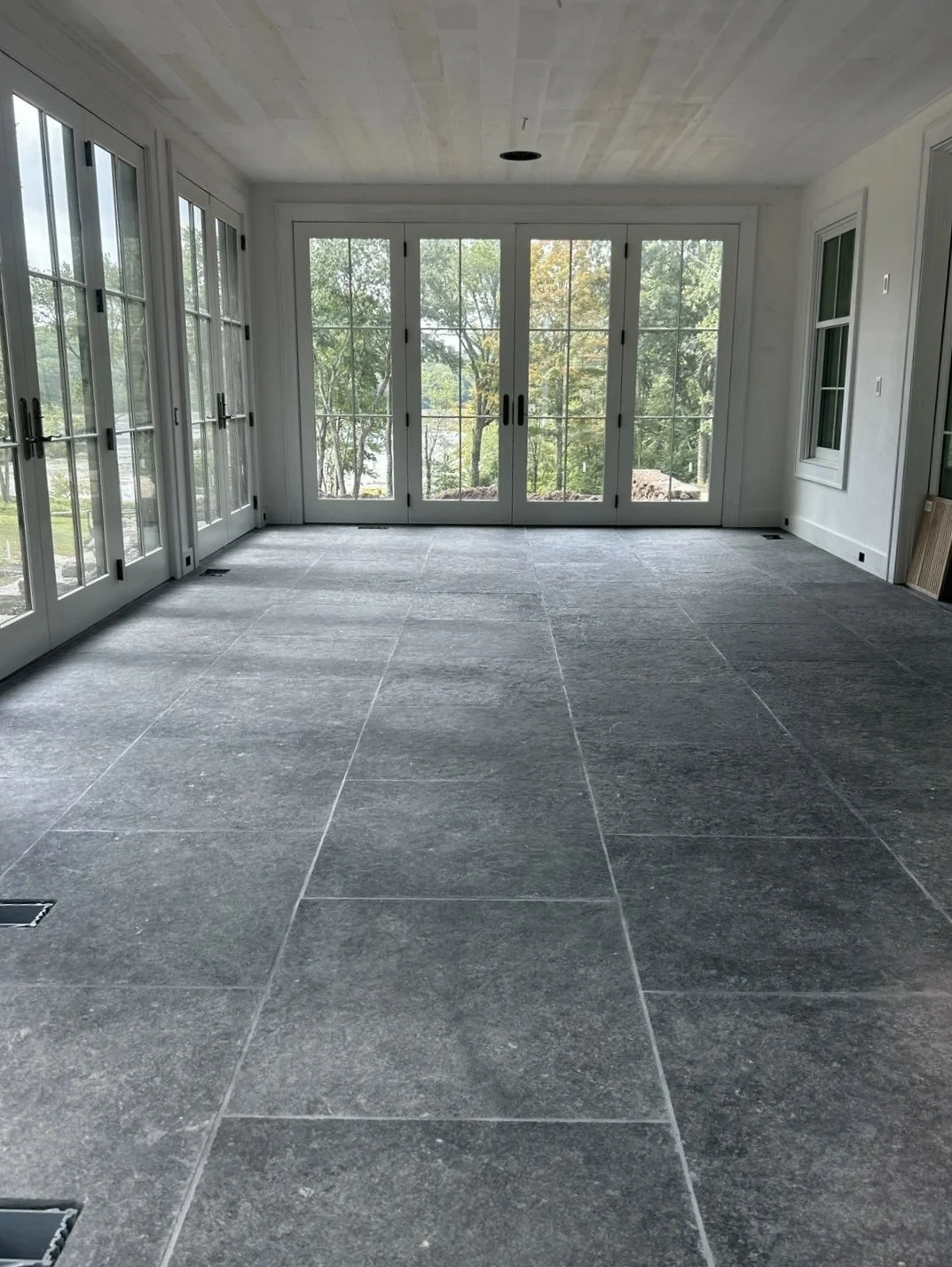 Empty sunroom with large glass doors and windows, gray tiled floor, and outdoor greenery visible through the glass.