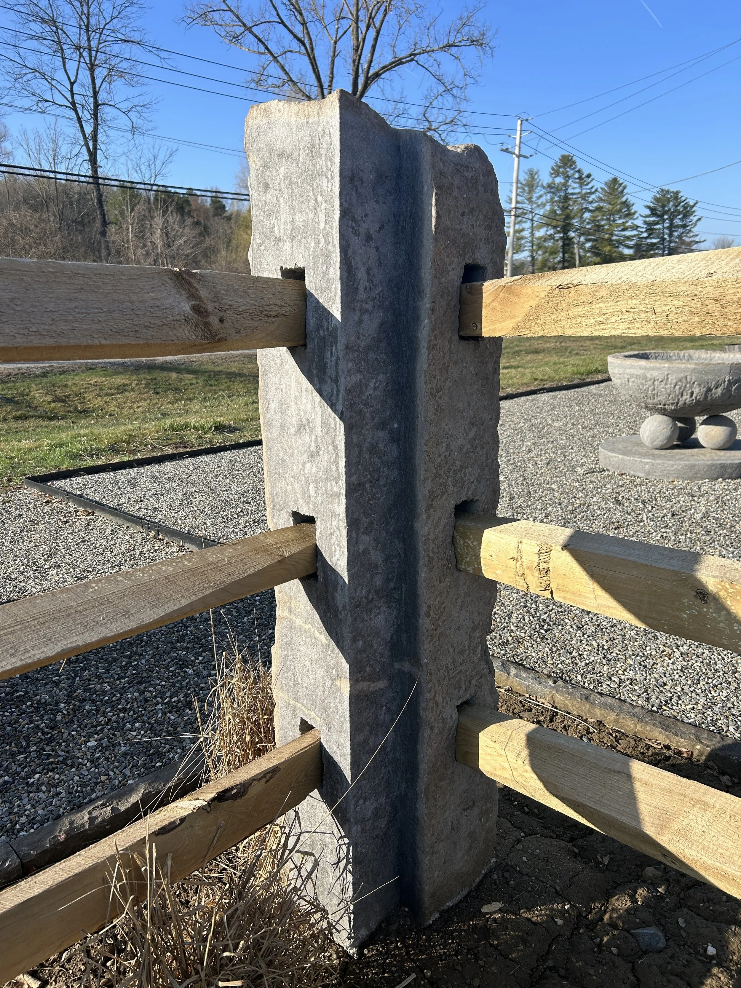 A concrete and wooden fence construction with a large concrete post and horizontal wooden beams attached with metal brackets in an outdoor setting with trees, power lines, and gravel ground.