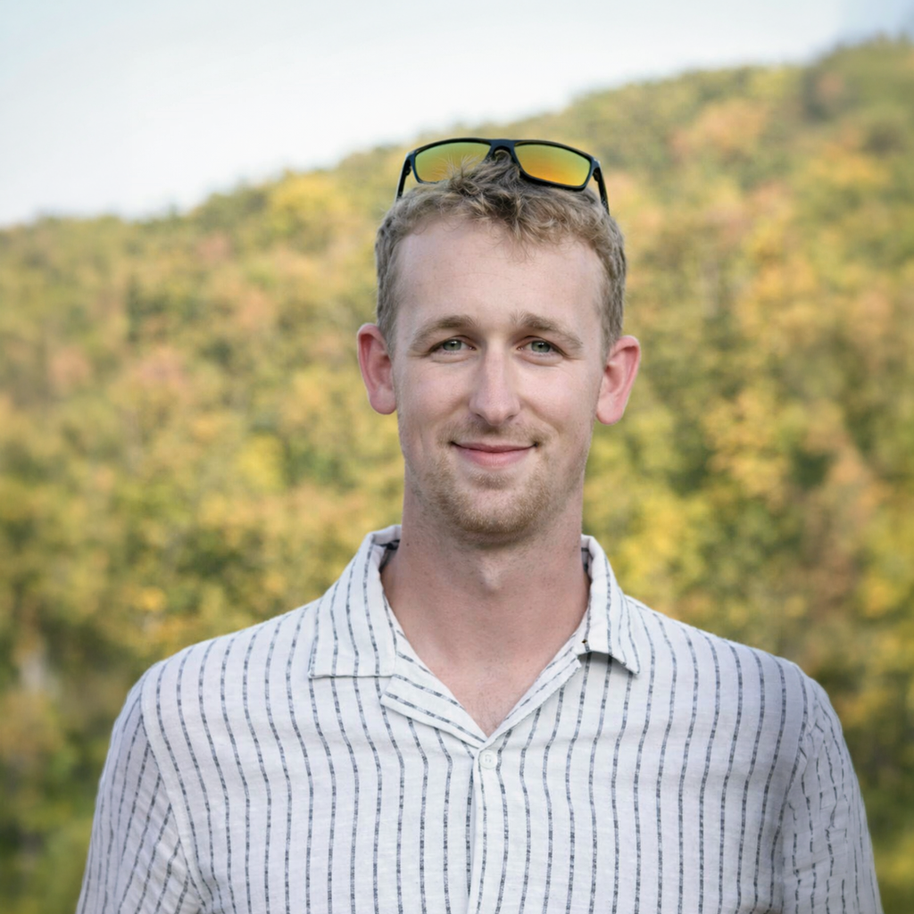 A man outdoors with sunglasses on his head, smiling, with a background of trees and hills during daytime.