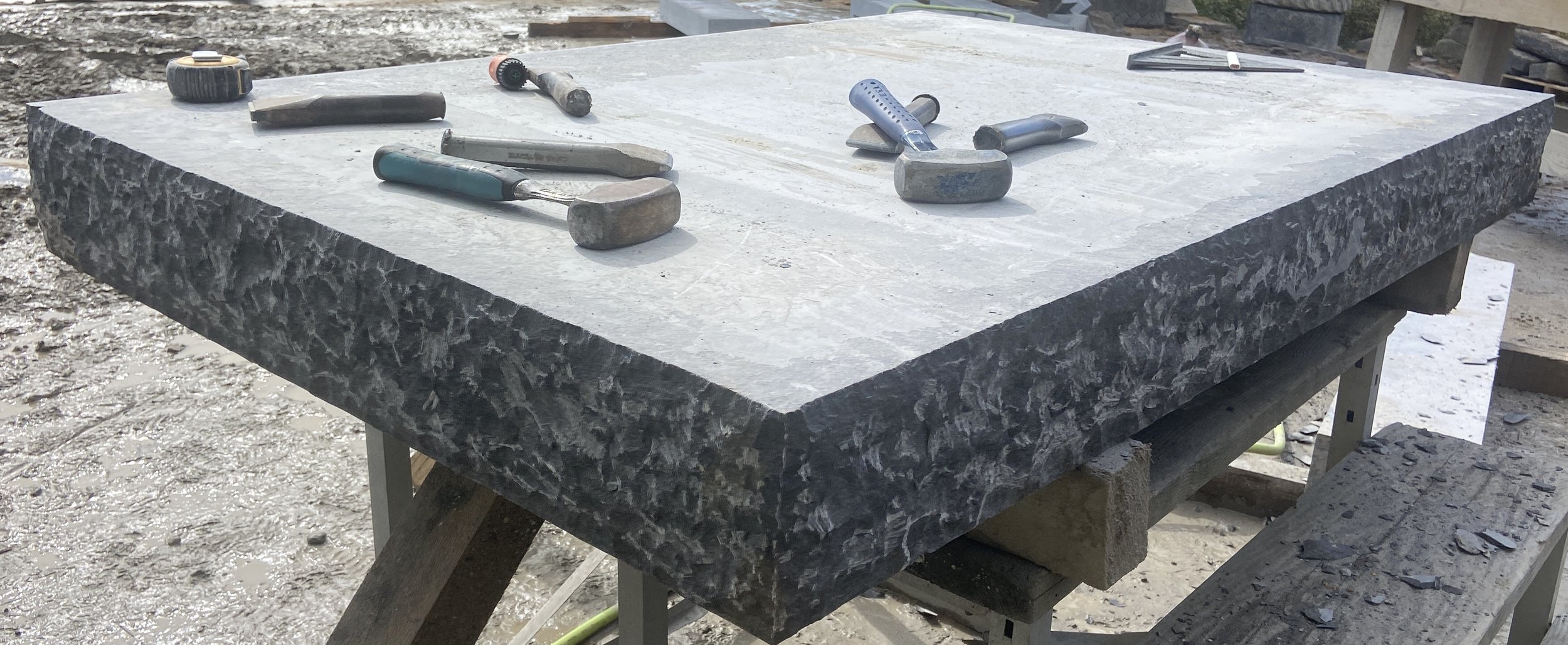 A large rectangular marble slab is resting on a wooden workbench, with several chisels and hammers placed on top of it. The marble has a gray veined pattern, and the background shows an outdoor construction or workshop area with dirt and tools.