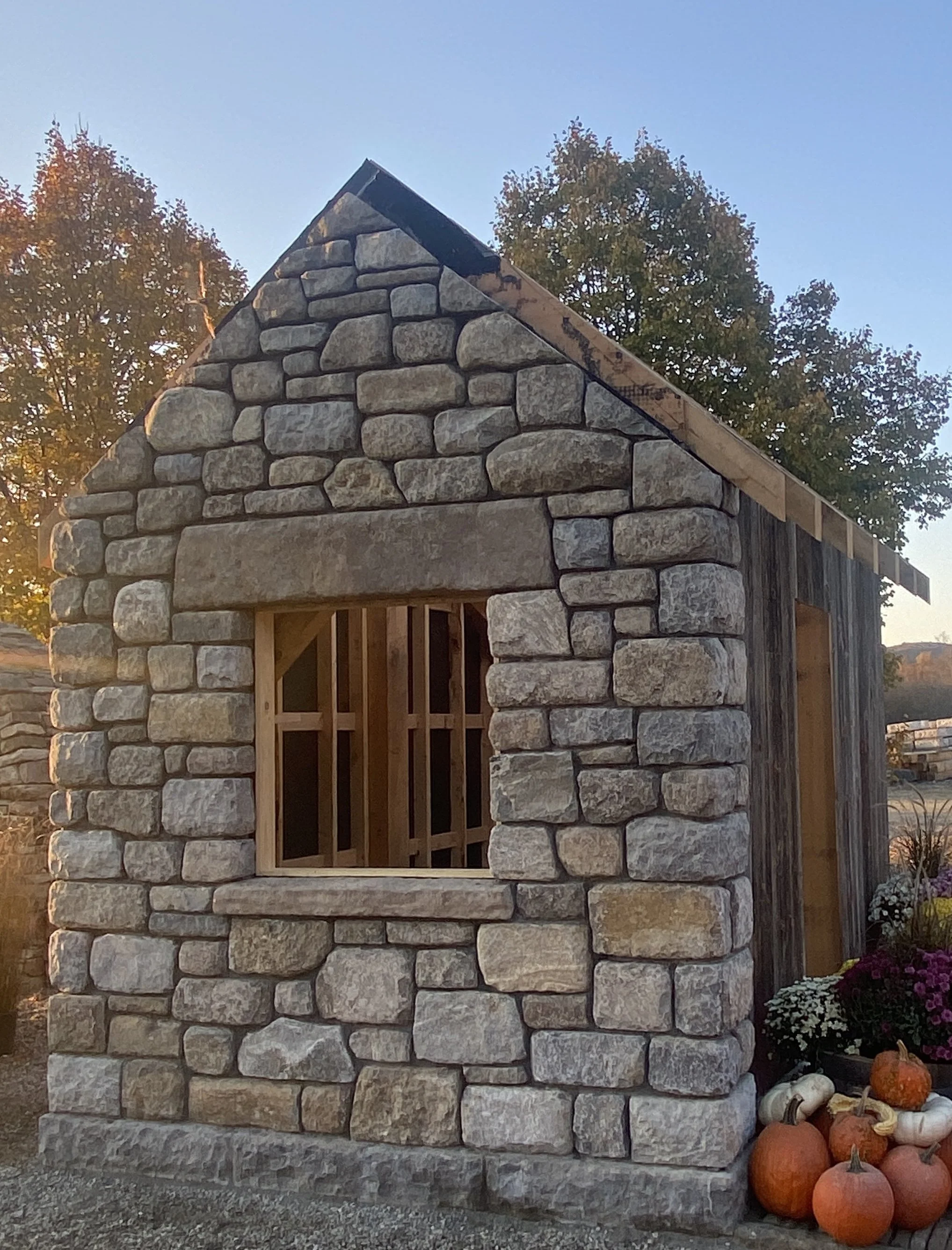 A small stone and wood shed with an unfinished window and roof, decorated with pumpkins and flowers outdoors during fall.