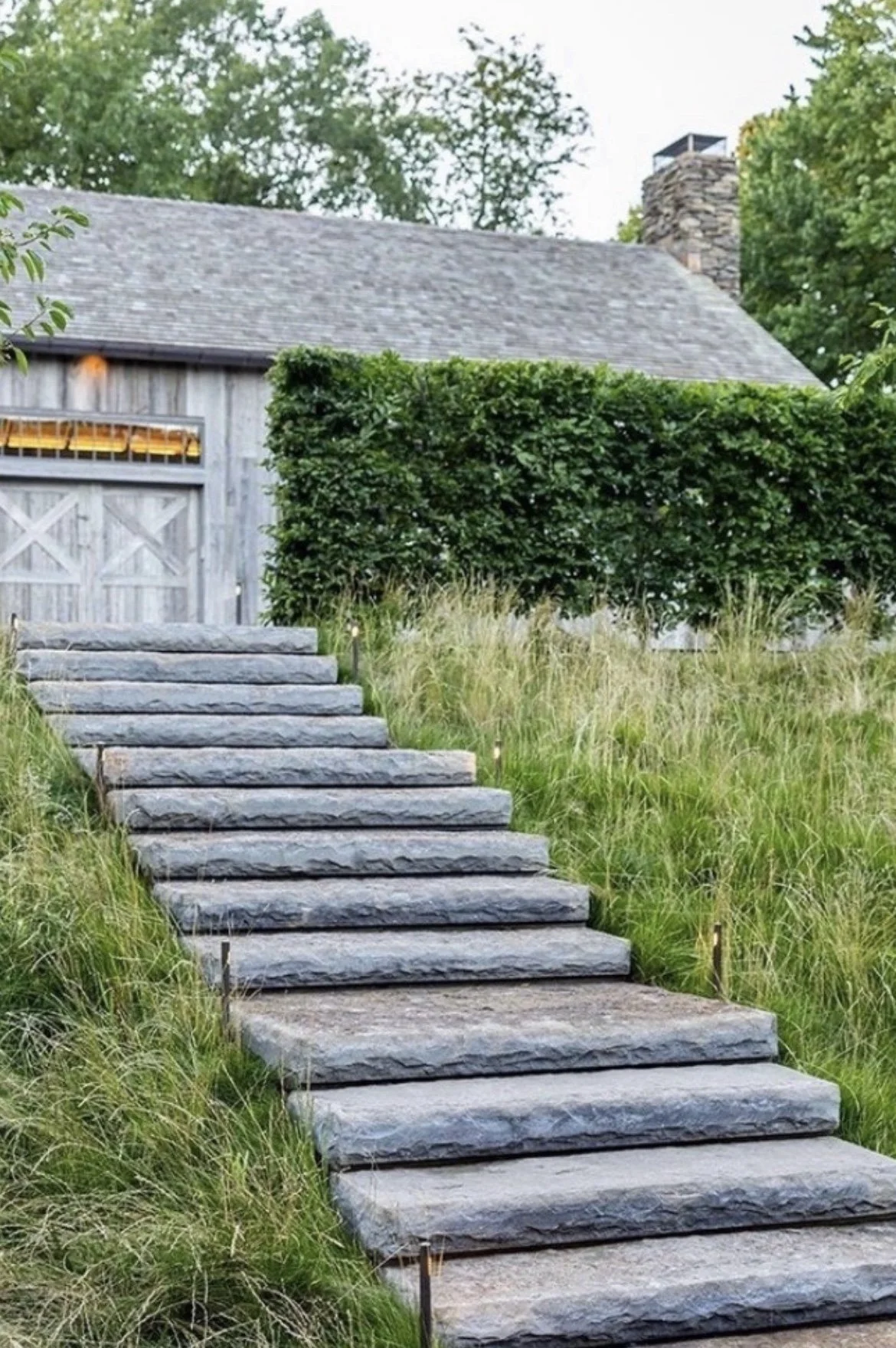 Stone pathway leading up to a house with a wooden gate, surrounded by green grass, shrubs, and trees.