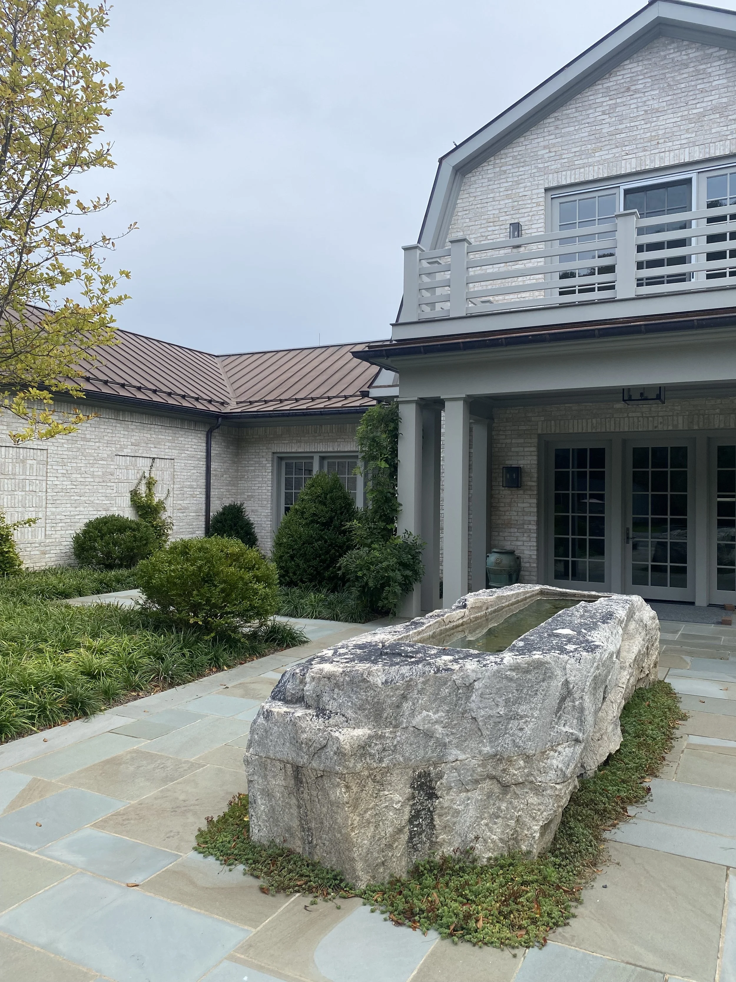 A large stone fountain outside a modern house with brick walls, a patio, and greenery.