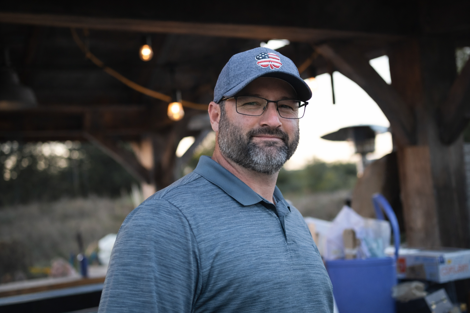A middle-aged man with glasses, a beard, and gray hair wearing a gray polo shirt and a gray cap with an American flag and paw print emblem, standing outdoors near a wooden structure with hanging lights.