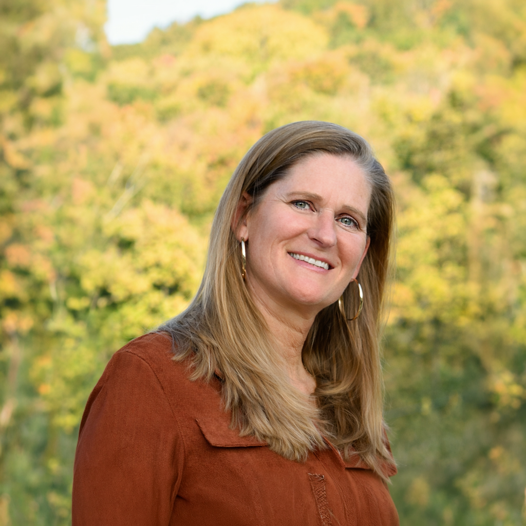A woman with long, blonde hair and hoop earrings smiling outdoors with a blurry background of green and yellow trees.