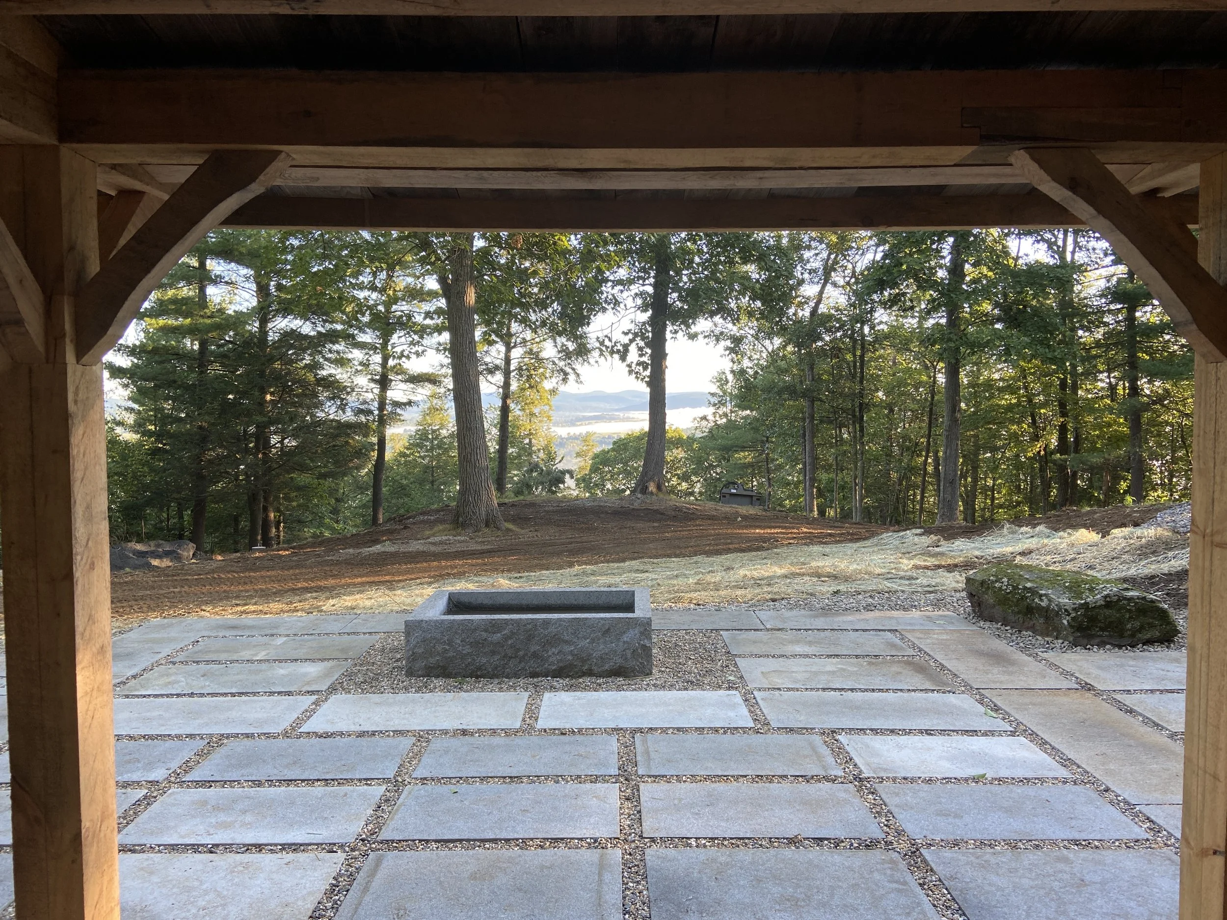 View from under a wooden structure looking out over a paved outdoor area with trees and hills in the distance.