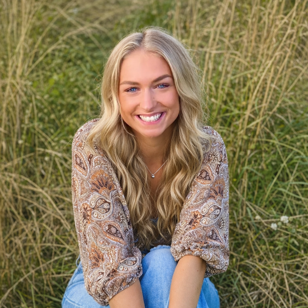 A young woman with long blonde wavy hair and blue eyes, smiling while sitting outdoors among tall grass.