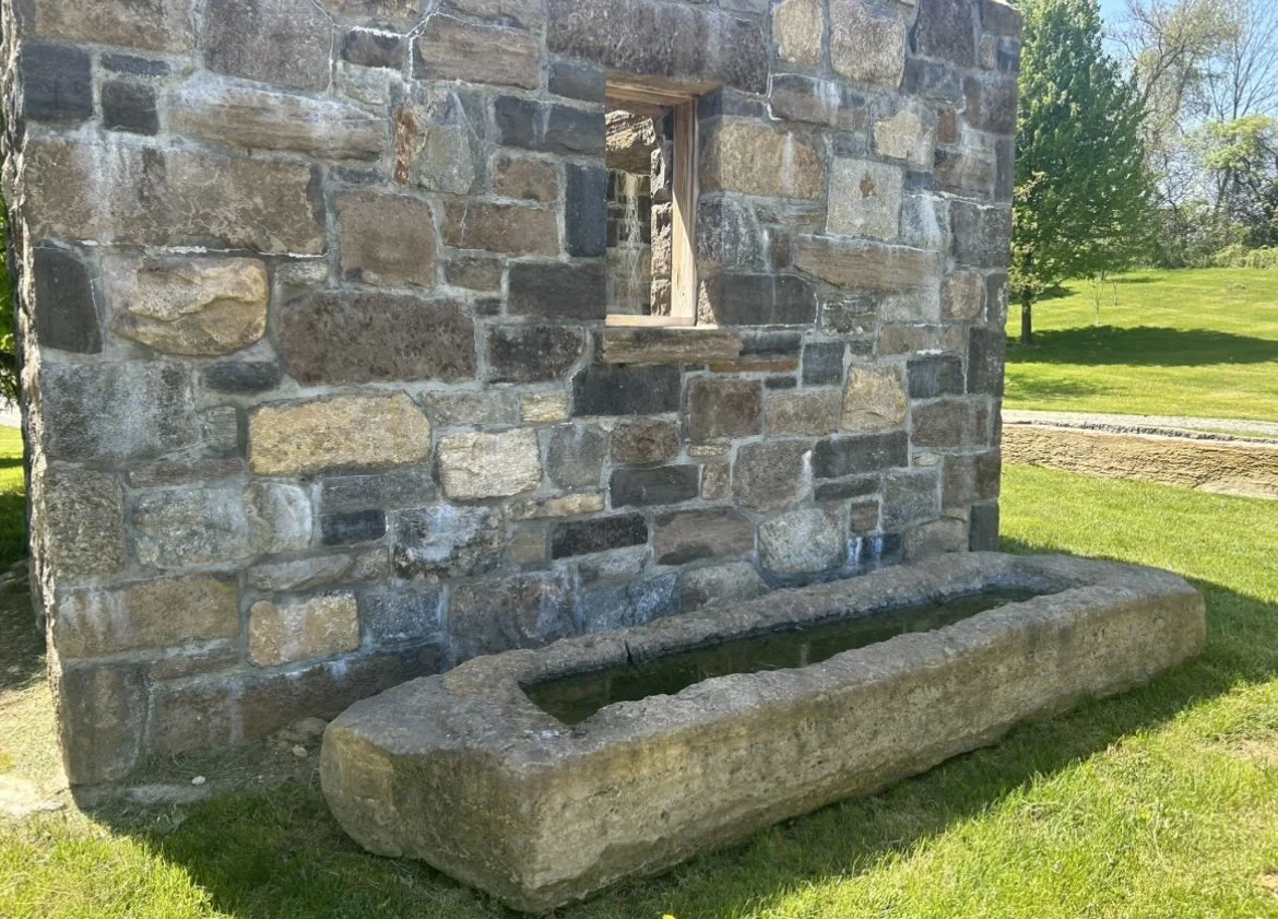 A stone building wall with a small rectangular window and a stone water trough on the grass outside.