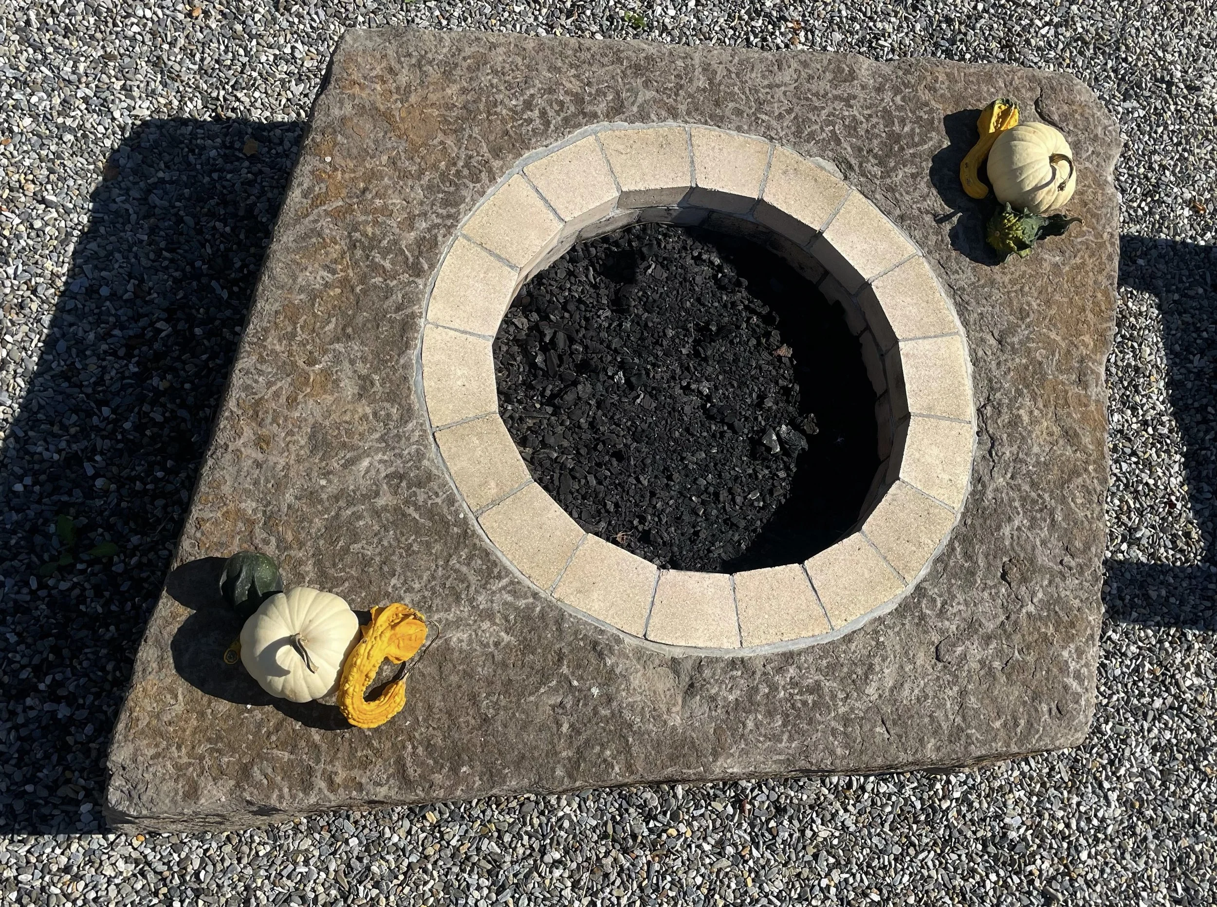 A square stone with a circular opening in the center, bordered by beige bricks, with small pumpkins and gourds on the corners, and surrounded by gravel.