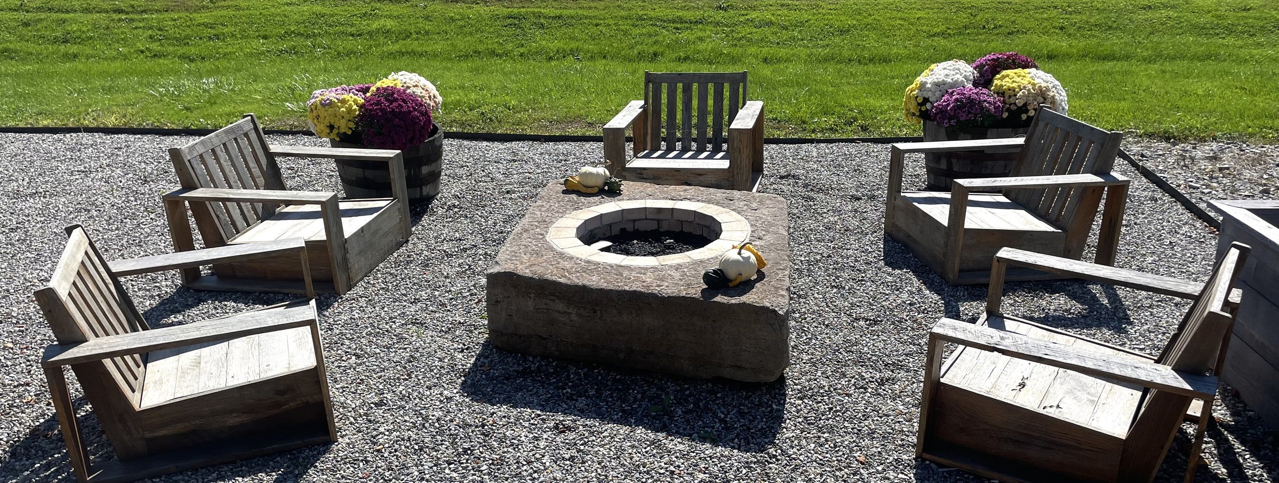 Outdoor fire pit surrounded by six wooden chairs on gravel with potted chrysanthemums in the background.