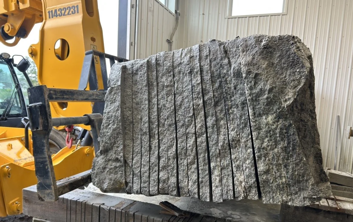 Large gray granite block with vertical and horizontal cut lines, secured on a wooden table inside a warehouse, with a yellow heavy machinery lift in the background.