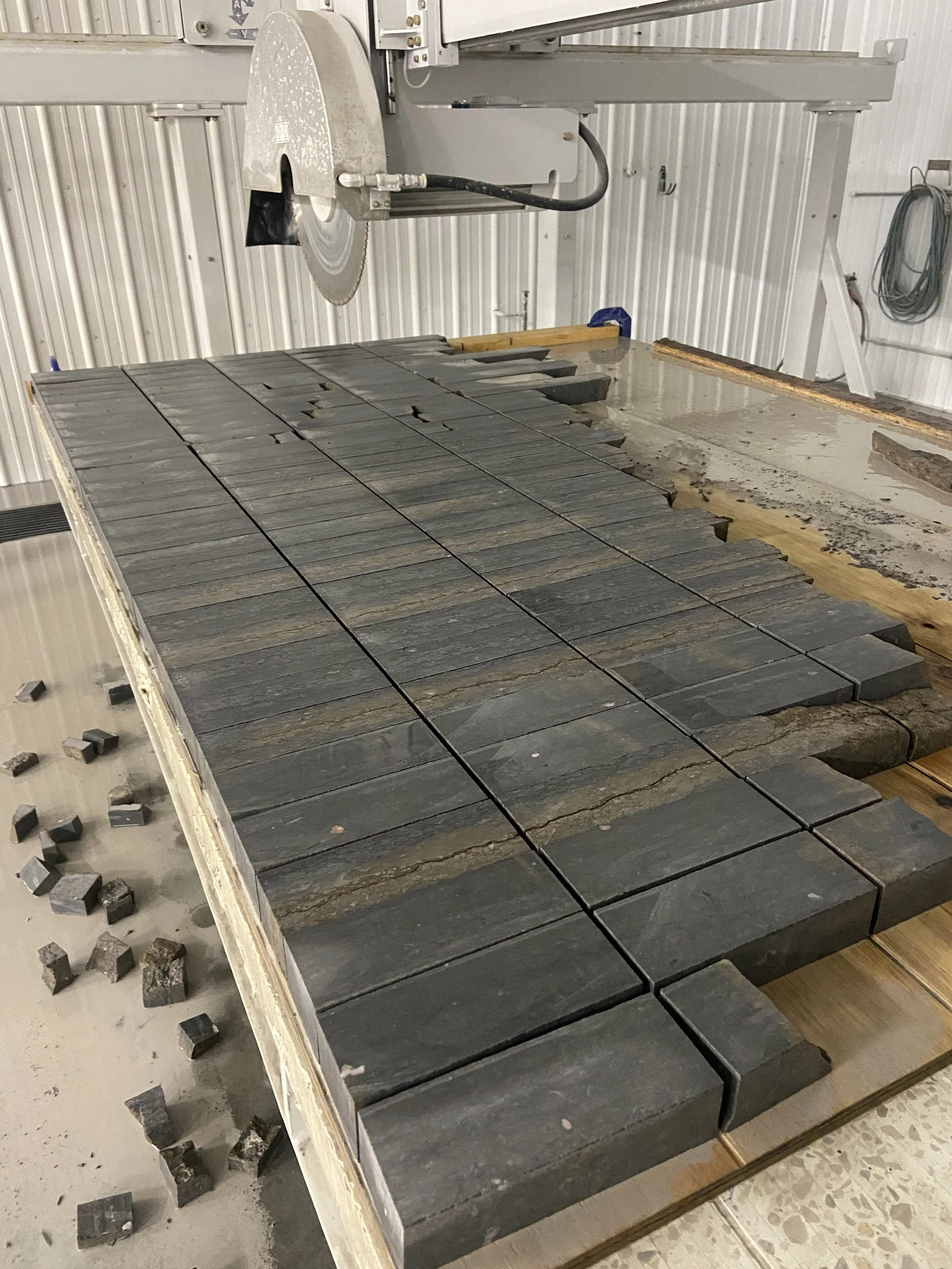 A saw cutting large black stone tiles, with some tiles already cut and arranged on a worktable in a workshop.