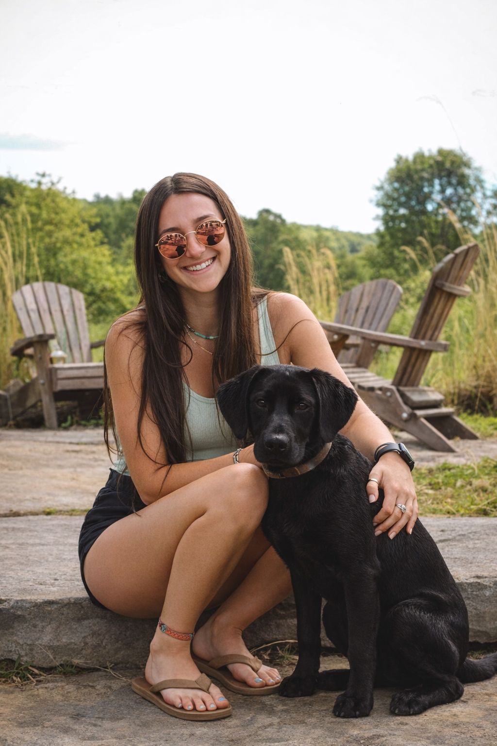 A woman with long brown hair wearing sunglasses, a light turquoise tank top, and black shorts sitting on a rock with a black Labrador puppy, outdoors in a natural setting with wooden Adirondack chairs and greenery in the background.