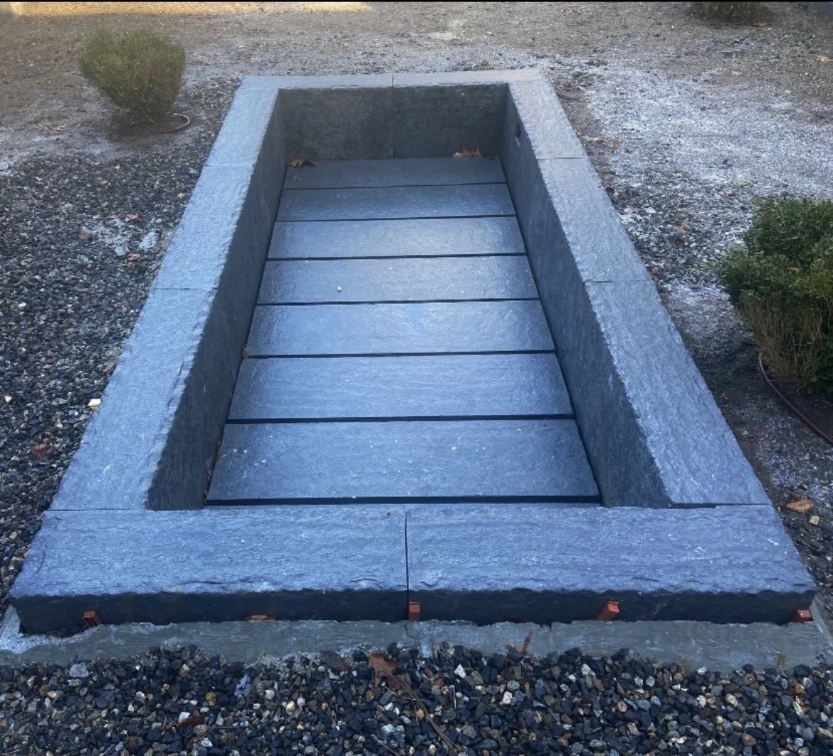 A rectangular stone fire pit with a wooden inner lining, located outdoors on gravel and dirt ground, with small bushes on either side.