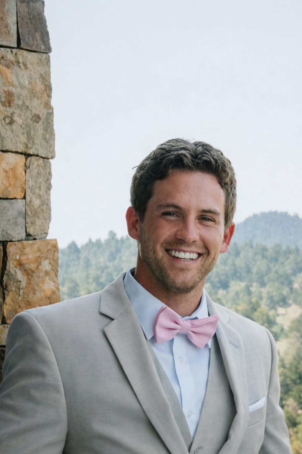 A smiling man in a light gray suit with a pink bow tie, standing outdoors near a stone wall with a scenic mountain and forest background.