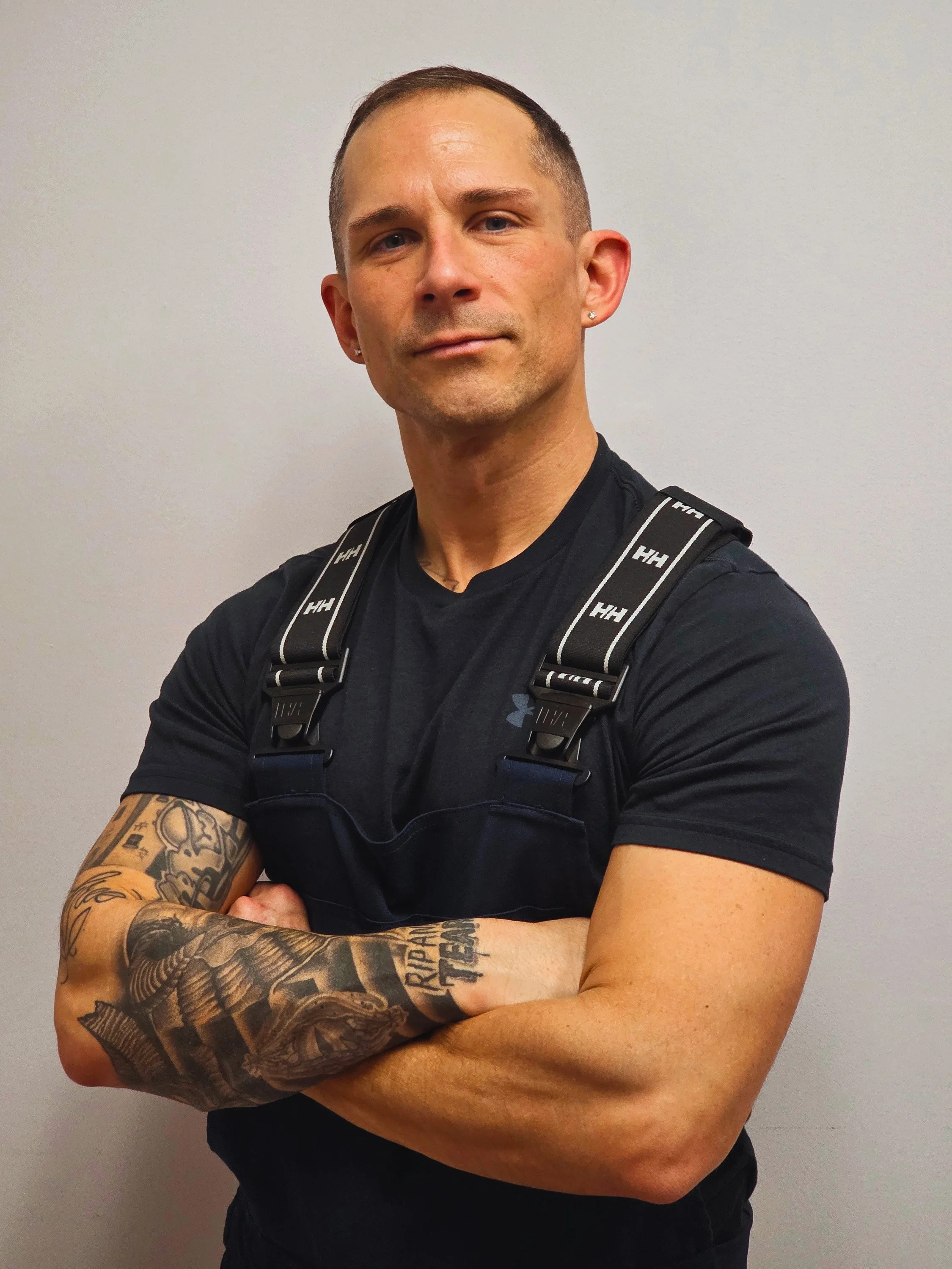 A man with short hair and earrings, wearing a black Under Armour shirt and navy blue coveralls, standing against a plain white wall with his arms crossed and looking at the camera.