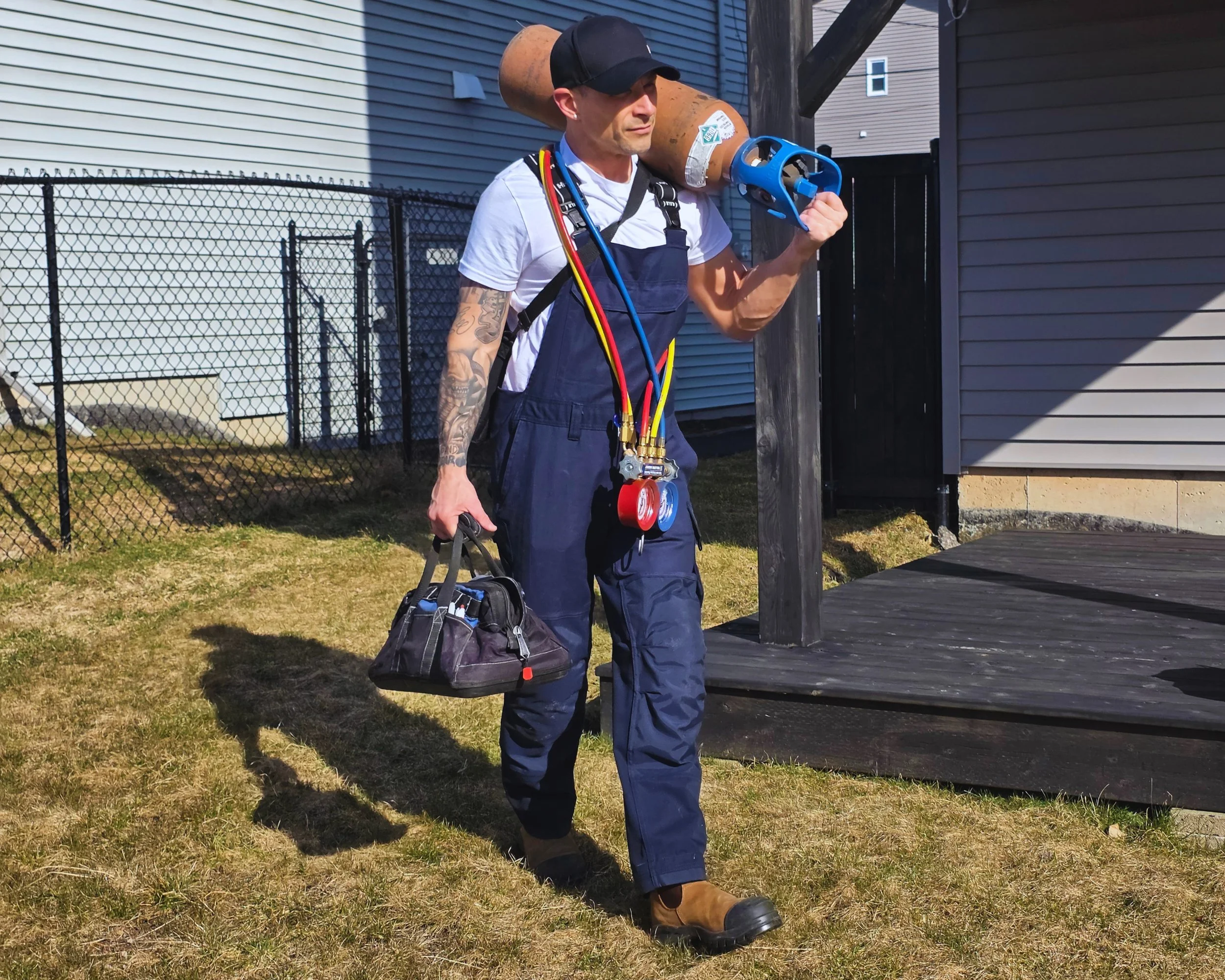 A man in work overalls stands outdoors carrying a gas line leak detection device on his shoulder and a toolbox in one hand, with refrigeration guages hanging from his neck.