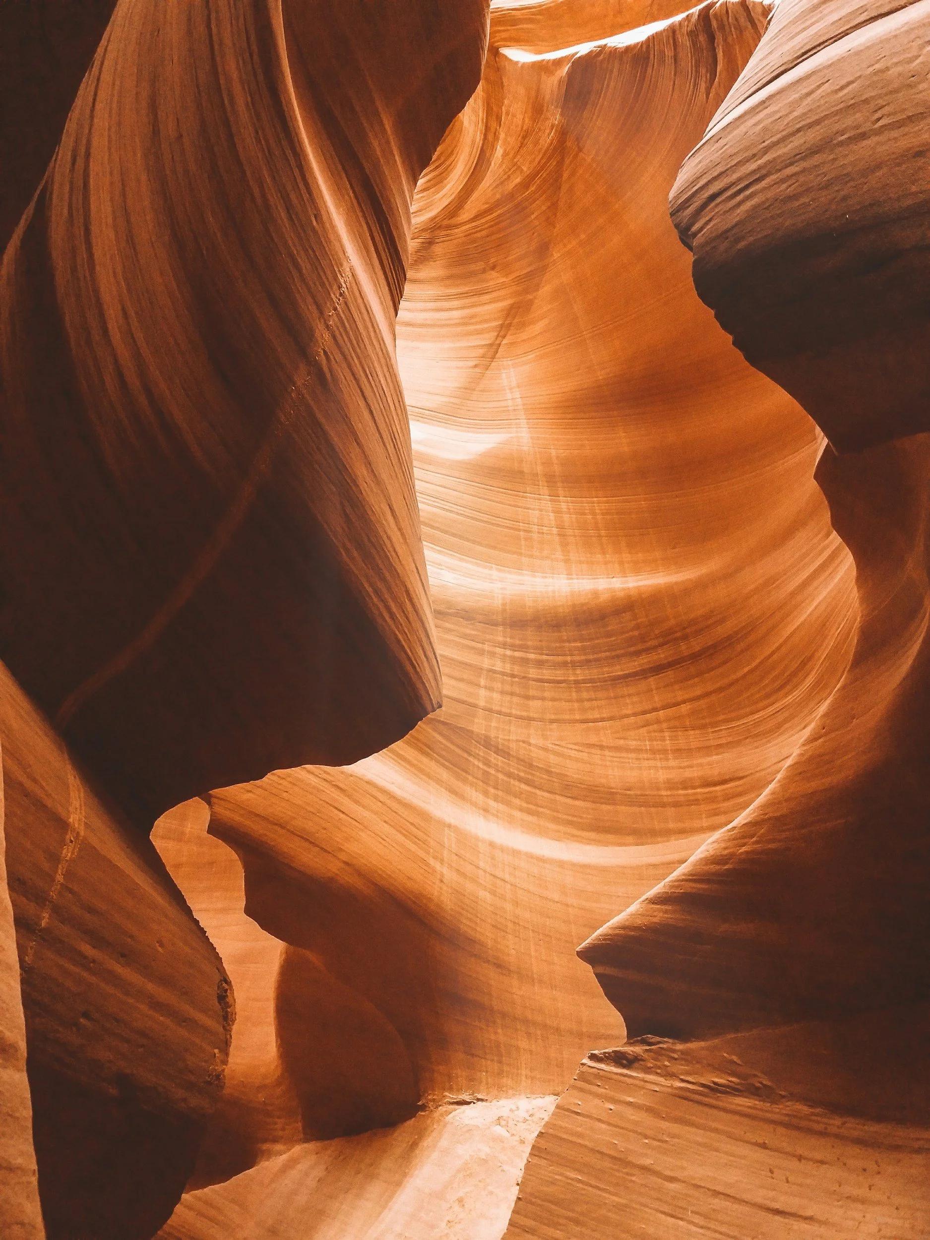A narrow slot canyon with smooth, flowing sandstone walls in shades of orange, brown, and tan, with light filtering down from above.