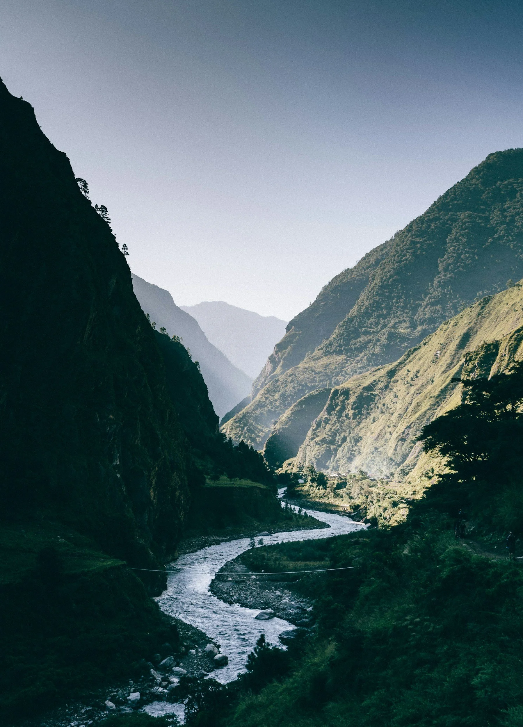 Scenic view of a river flowing through a lush green valley surrounded by towering mountains under a clear sky.