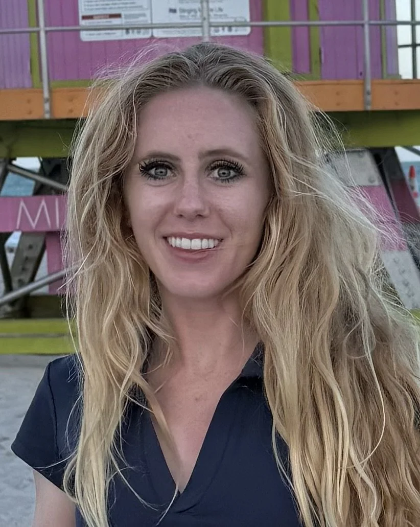 A woman with long, wavy blonde hair and blue eyes smiling at the camera, standing outdoors near a colorful structure with pink, purple, and green panels.
