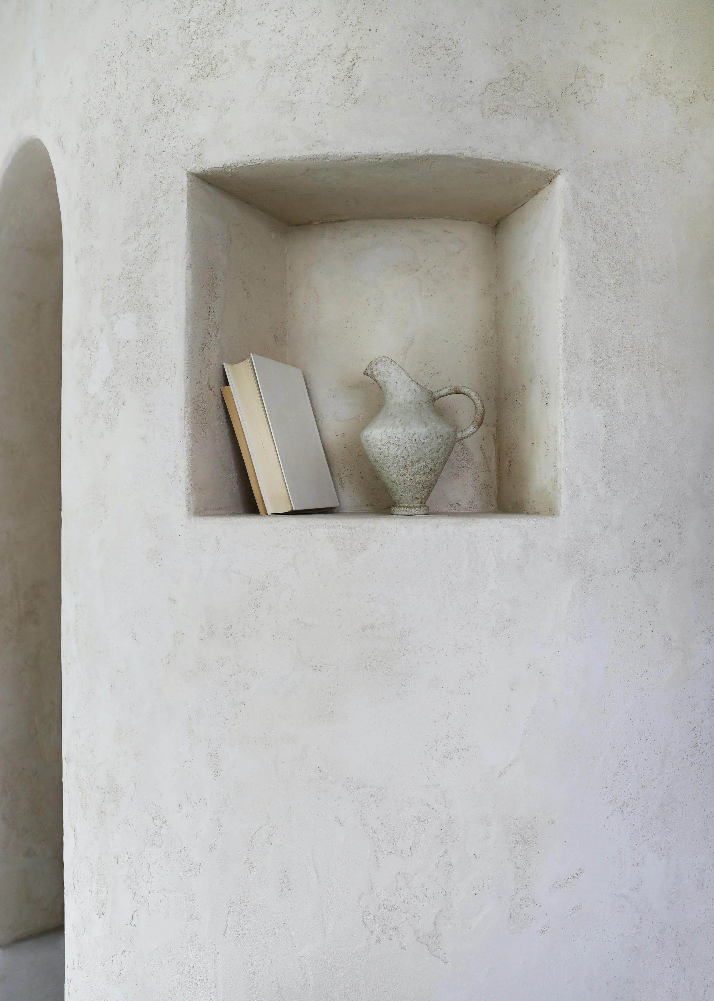 A recessed wall shelf holding two books and a ceramic pitcher against a textured, white stucco wall.