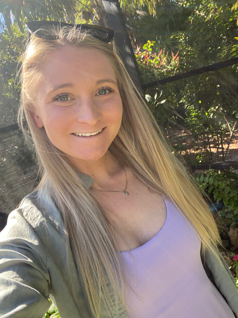 A smiling young woman with long blonde hair and sunglasses on her head, wearing a light purple top and a gray jacket, standing outdoors with greenery and sunlight in the background.
