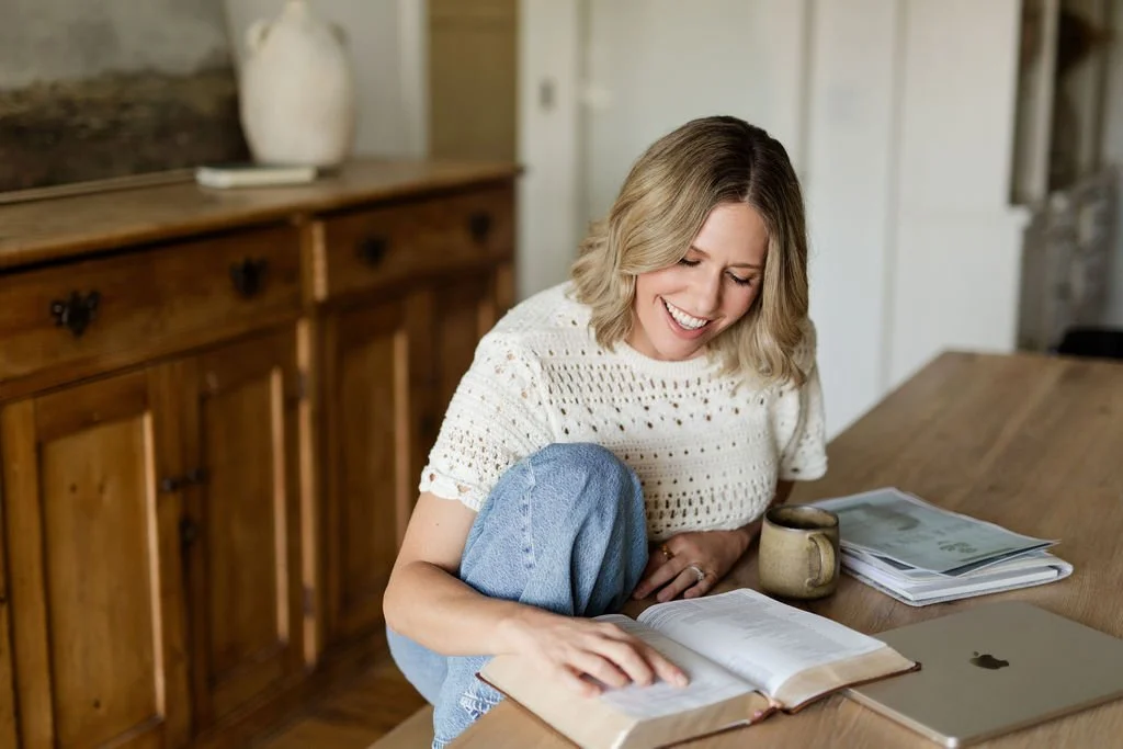A woman with shoulder-length blonde hair, wearing a white knit top, is sitting at a wooden table, smiling, and reading a book. There is a laptop, a mug, and some papers or magazines on the table. The background shows a wooden cabinet and a cozy, well-lit room.
