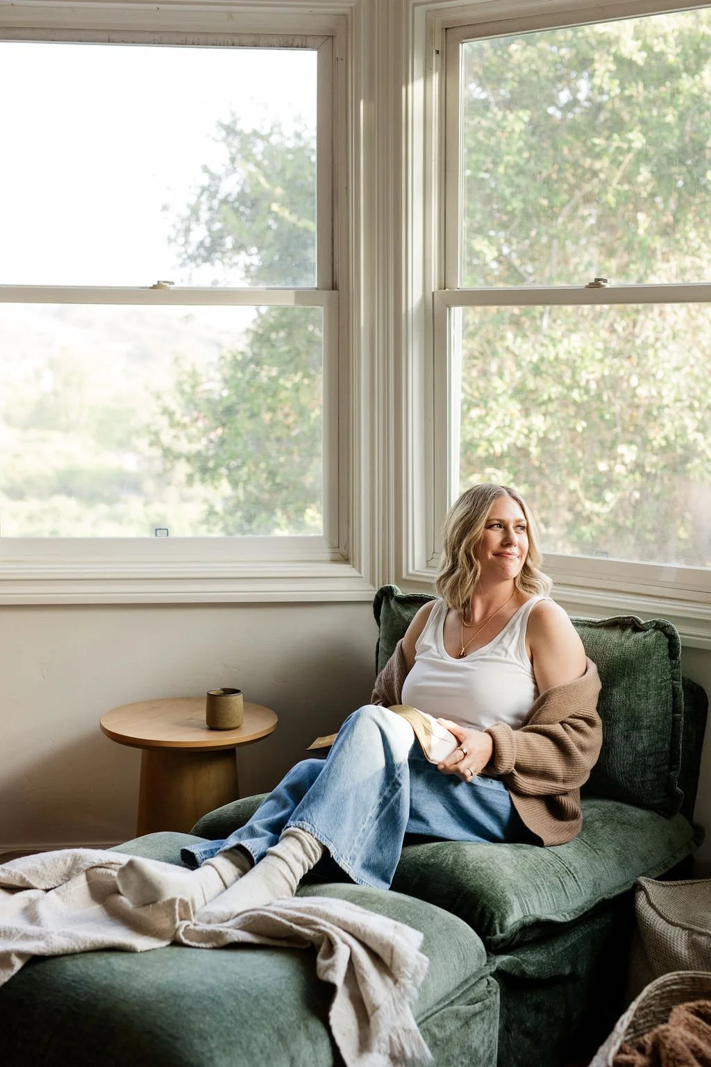 A woman with blonde hair sitting on a dark green sofa near large windows with trees outside, holding a book, dressed in a white sleeveless top, light blue jeans, a brown cardigan, and socks.