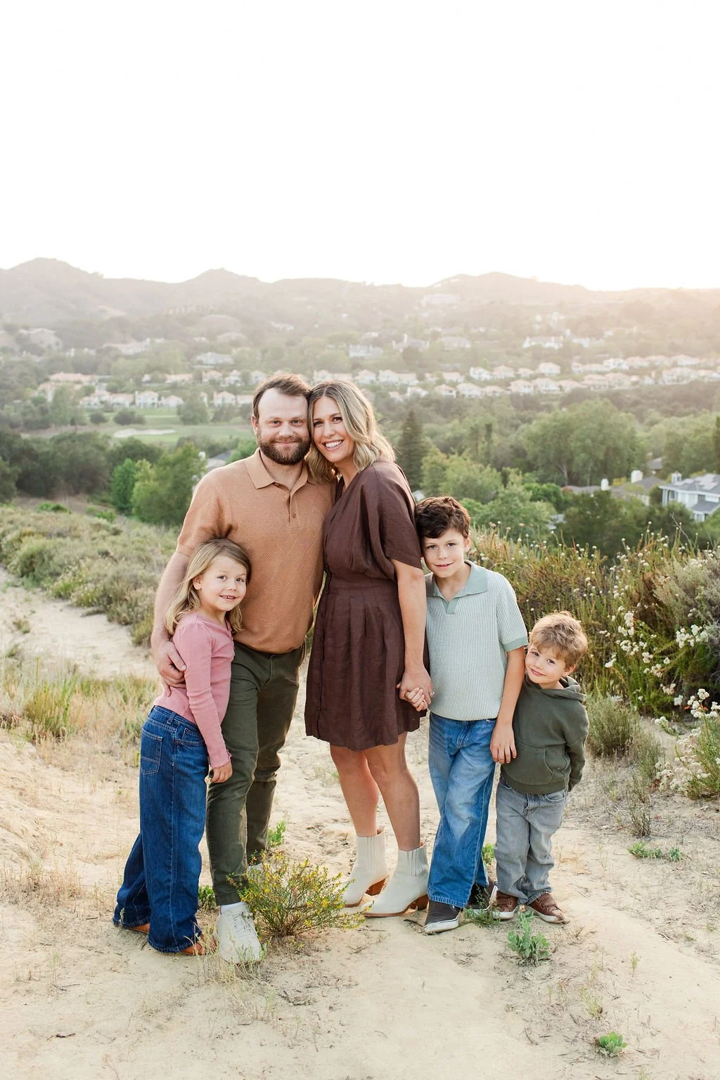 A family of five, including a man, woman, and three children, standing outdoors on a dirt path with hills and houses in the background, during sunset.