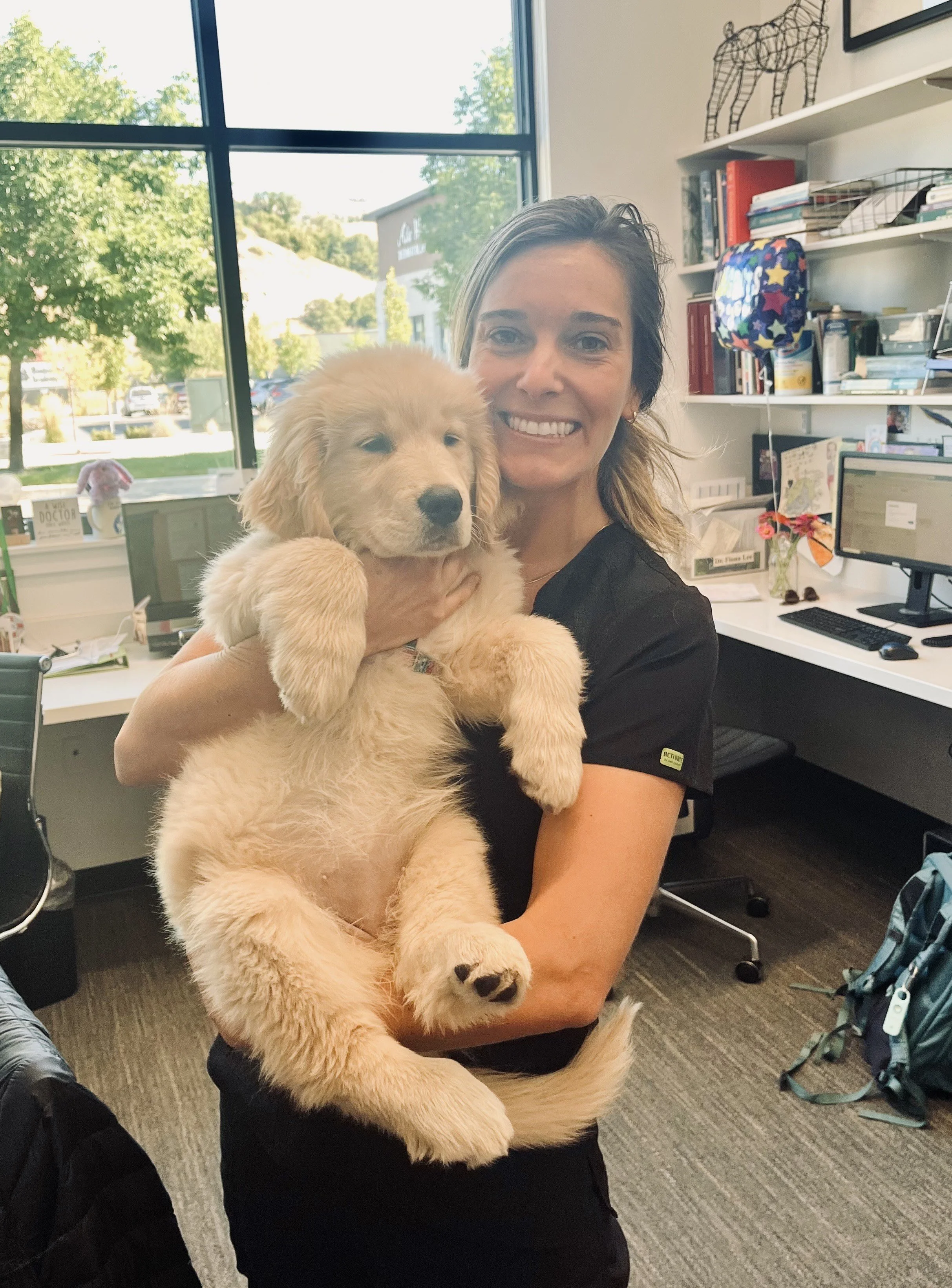 A woman smiling and holding a golden retriever puppy inside an office with large windows, bookshelves, and computer monitors.