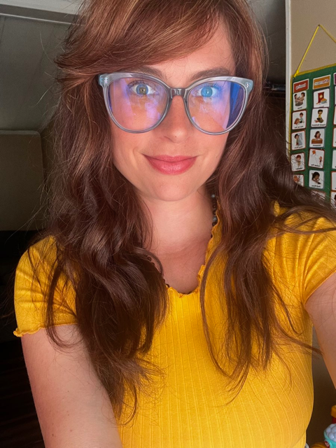 Woman with long wavy red hair wearing large clear glasses with blue light reflection, smiling, in a yellow shirt, in front of a colorful educational poster.