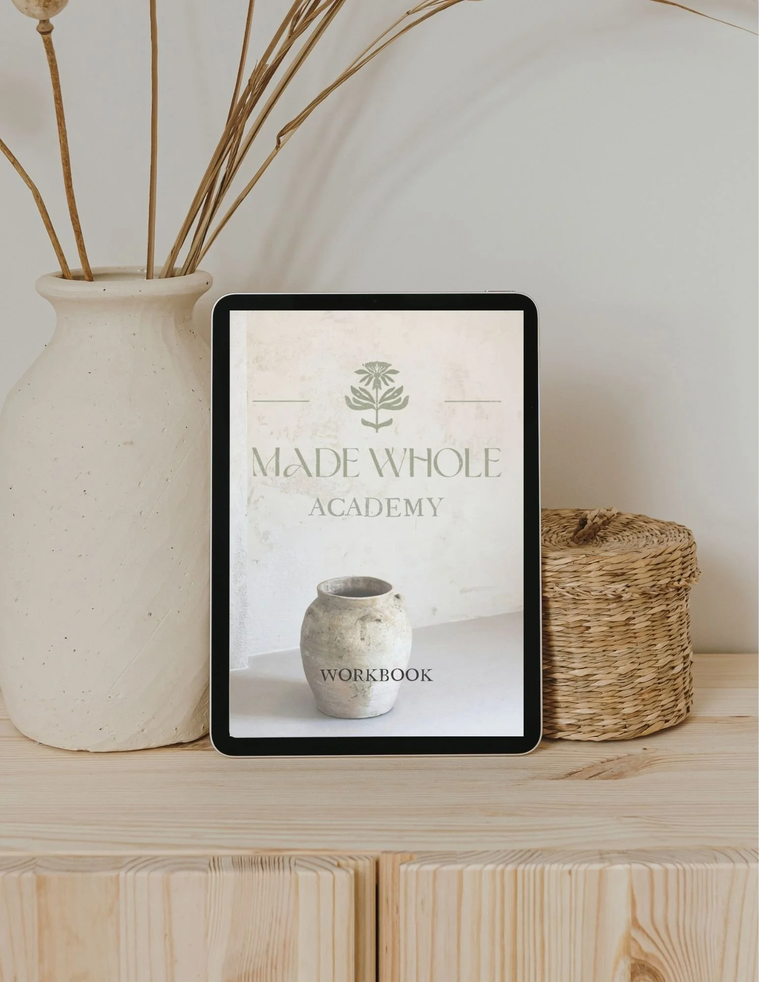 A tablet displaying a workbook titled 'Made Whole Academy' with an image of a white pottery vase on a light background. The tablet is placed on a wooden surface next to a large white ceramic vase with dried plant stems inside. To the right of the tablet, there is a woven basket. The background consists of a white wall.