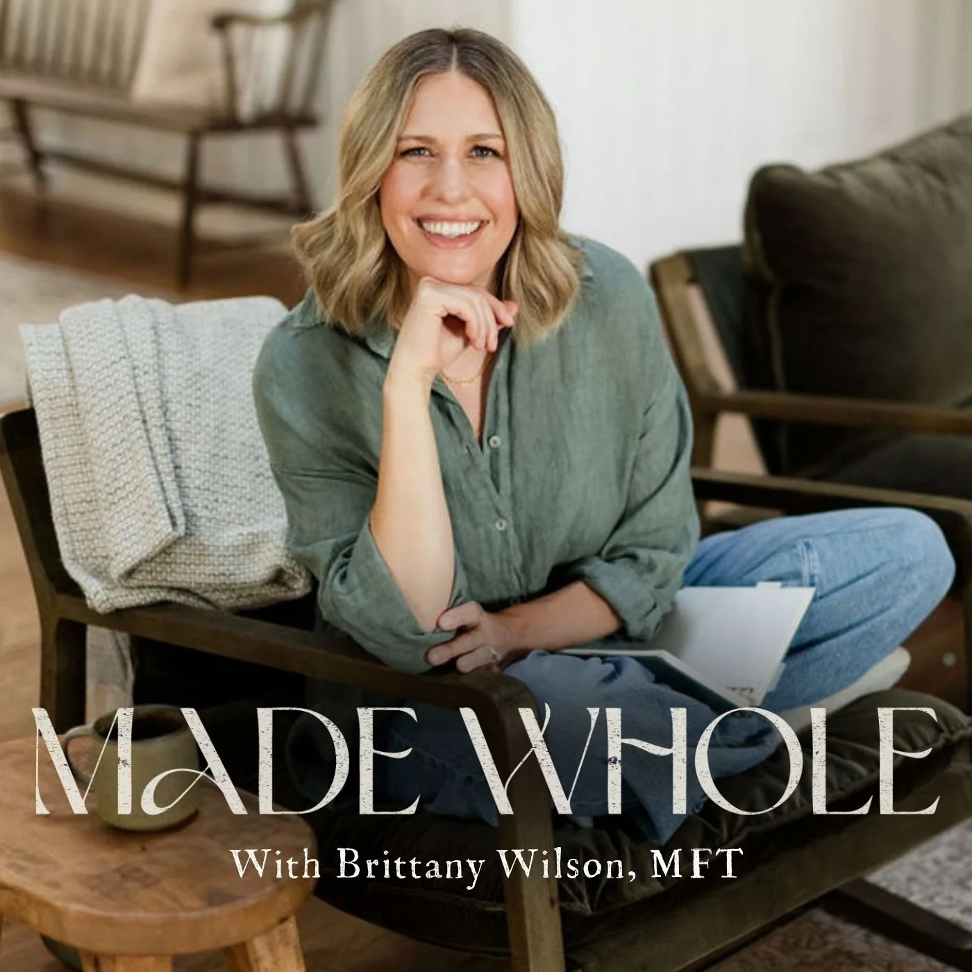 A woman with shoulder-length blonde hair, wearing a green shirt and blue jeans, sitting on a wooden chair in a cozy room with light-colored walls. She is smiling, resting her chin on her hand, and has a book in her lap. In the background, there is a bookshelf and a sofa. The text overlay reads: "MADE WHOLE With Brittany Wilson, MFT."