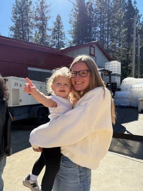 A smiling woman with long hair wearing glasses and a cream sweater, holding a young girl with curly blonde hair in a white shirt outdoors on a sunny day.