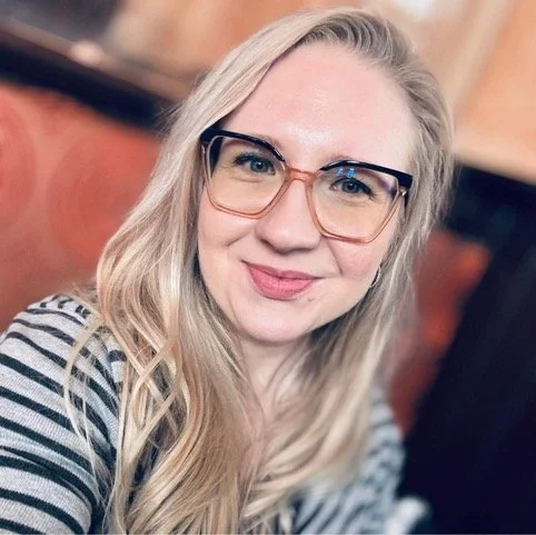 A young woman with long blonde hair wearing large glasses and a black and white striped shirt, smiling at the camera.