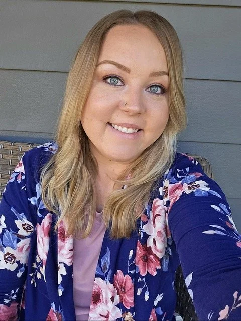 A woman with long blond hair and blue eyes taking a selfie outside, wearing a floral jacket over a purple top, smiling.