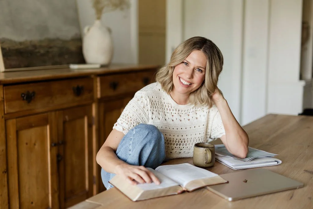 A woman with blonde hair wearing a white knit top sits at a wooden table, smiling at the camera. She has an open book, a laptop, a cup, and some papers in front of her.