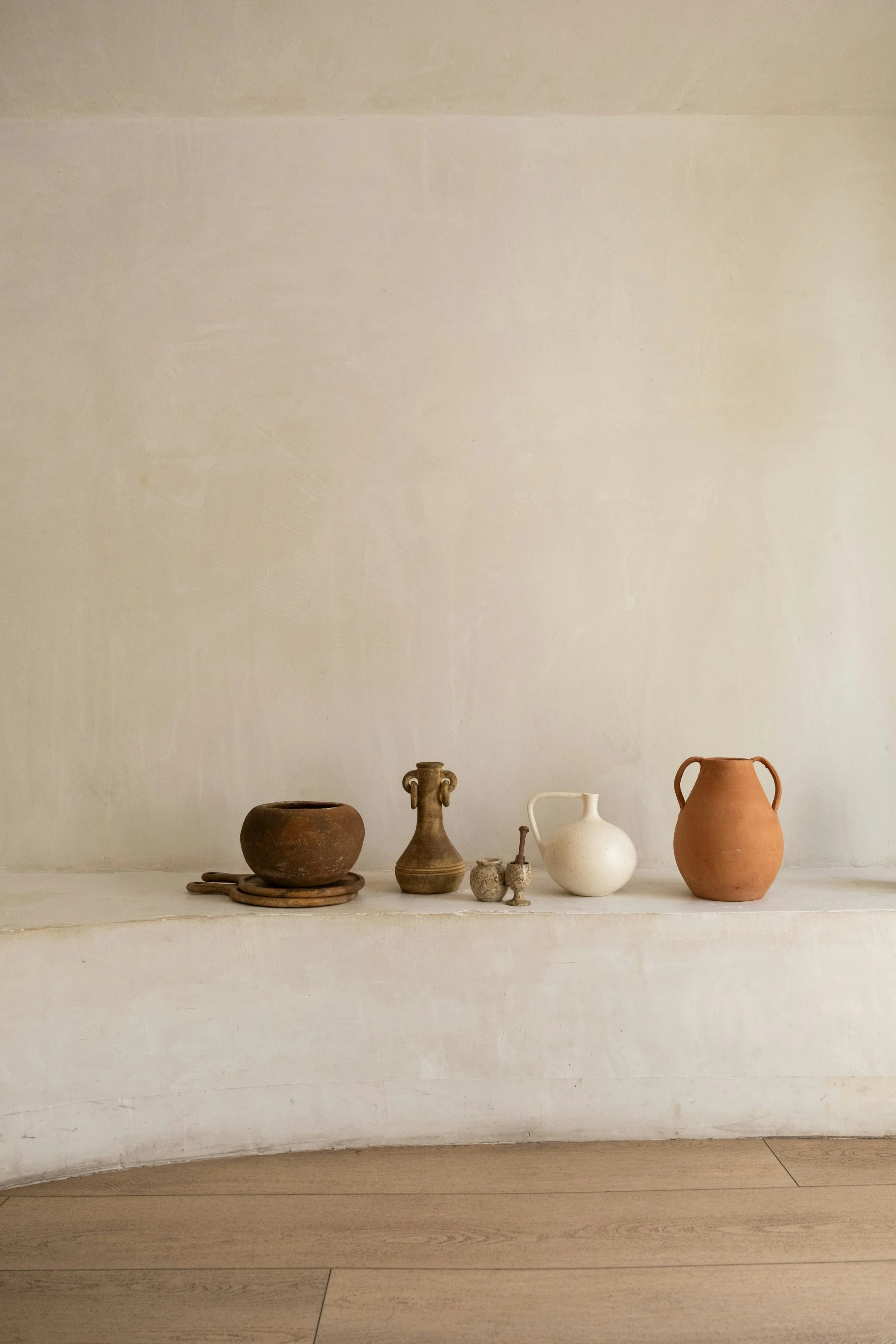 Various pottery vessels, including bowls and vases, displayed on a white shelf against a plain wall with a wooden floor below.