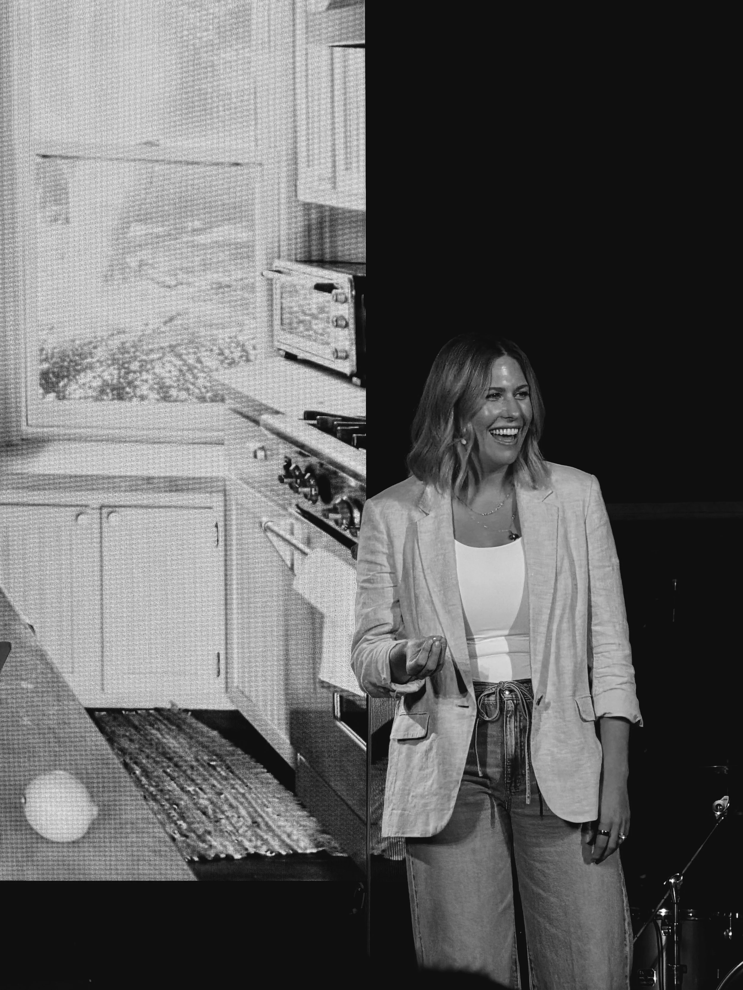 A woman with shoulder-length hair smiling and wearing a blazer and jeans, standing on stage with a large screen behind her displaying an outdoor scene with a stove and trees, in black and white.