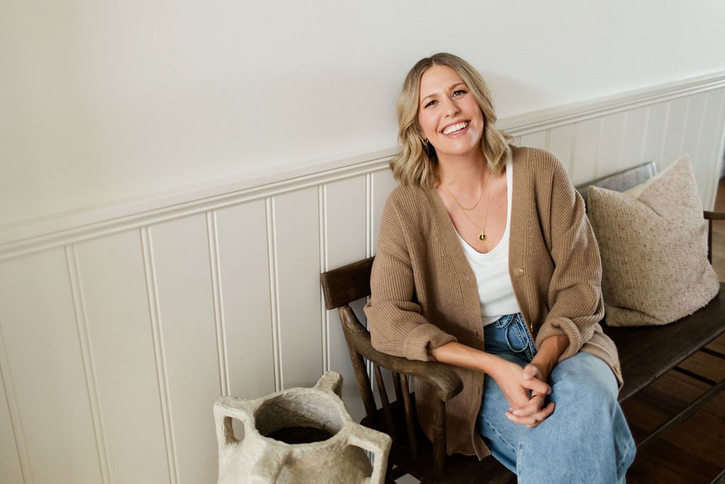 A woman with blonde hair, wearing a tan cardigan, white t-shirt, and blue jeans, smiling while sitting on a wooden bench with cushions, in a room with white paneling on the wall.