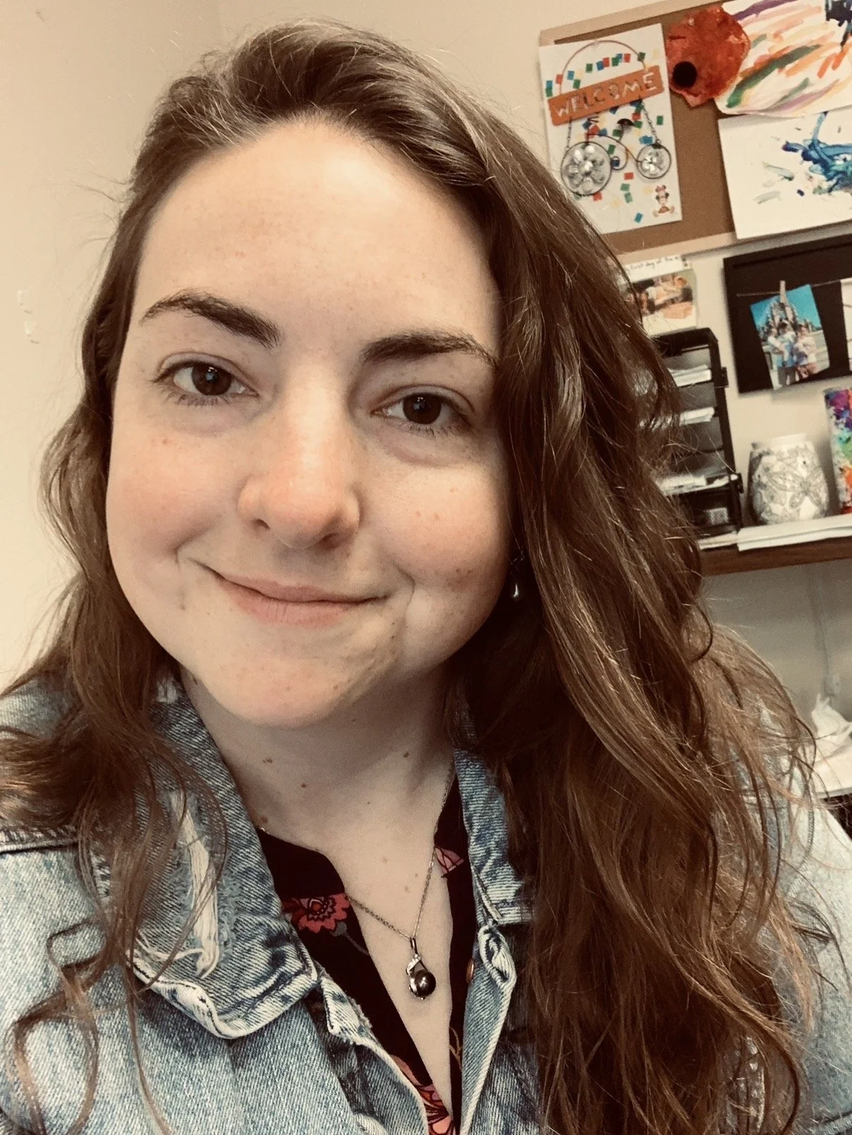 A young woman with long curly brown hair and light skin, smiling slightly, wearing a denim jacket over a dark shirt with red patterns, against a background of a bulletin board and colorful artworks.